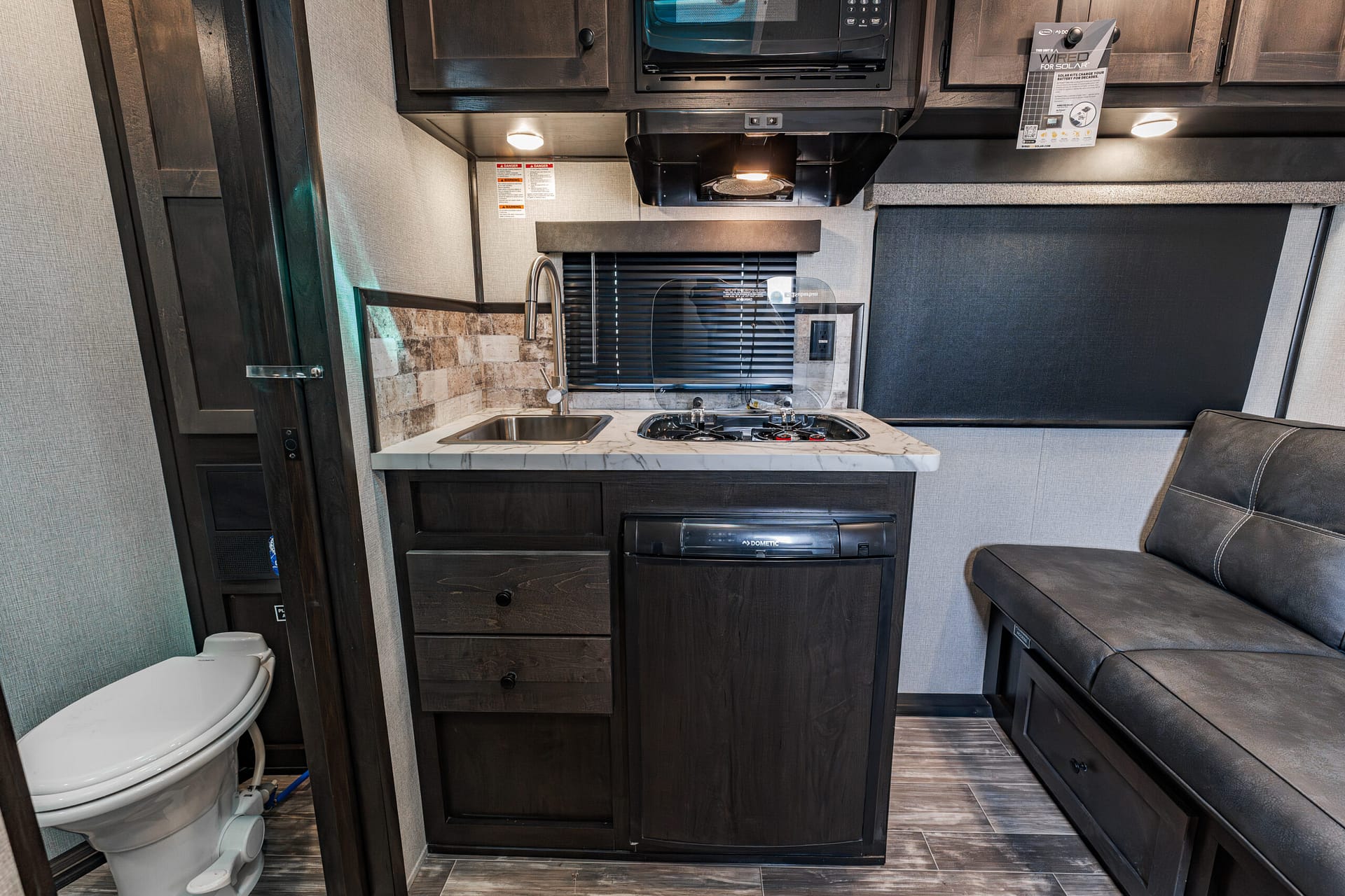 Compact RV kitchen with sink, two-burner stove, marble countertop, and dark wood cabinets.