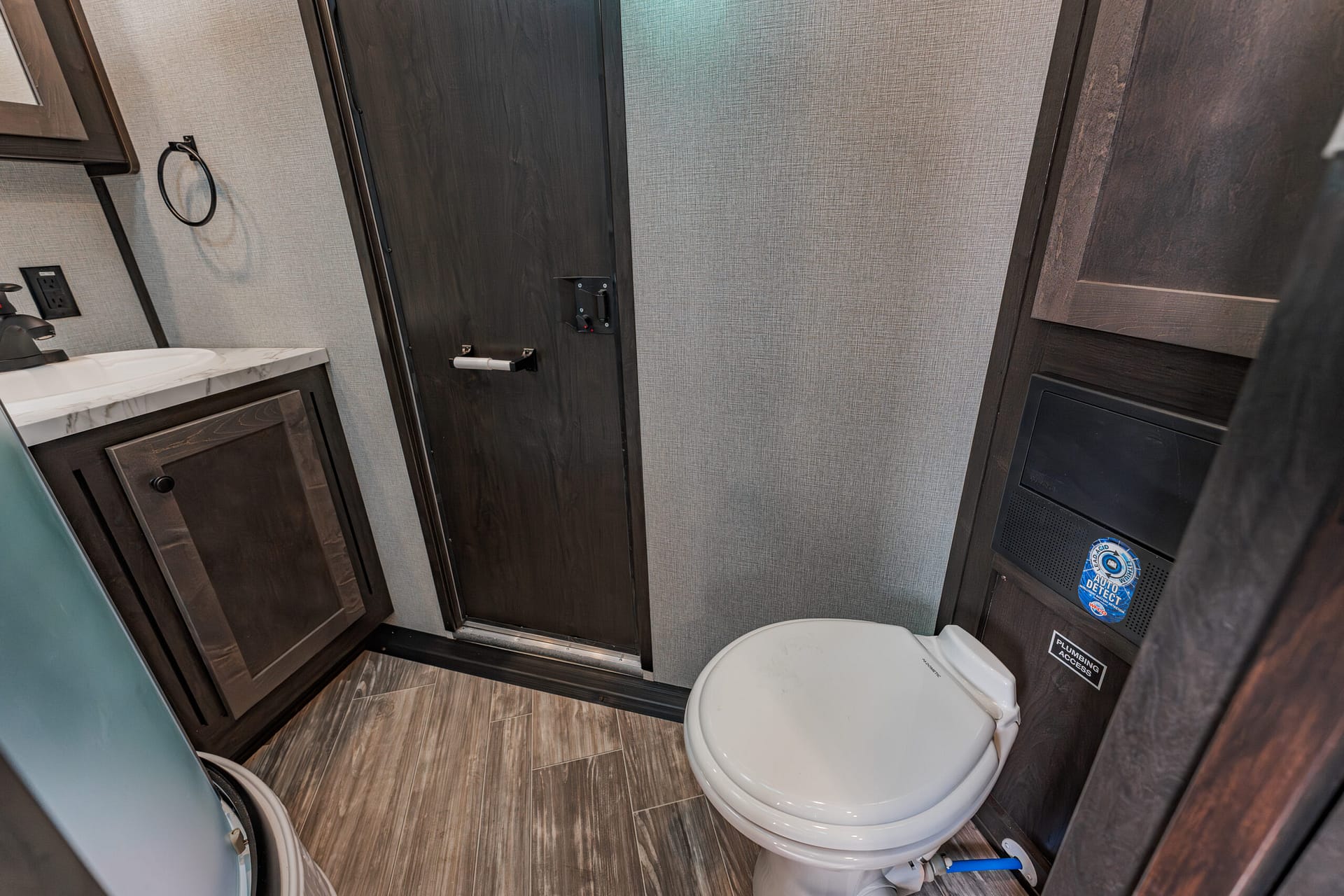 Small bathroom in a trailer: white toilet in the foreground, dark wood cabinets, and a marble-topped sink on the left with a towel ring nearby. A door is centered in the back wall.