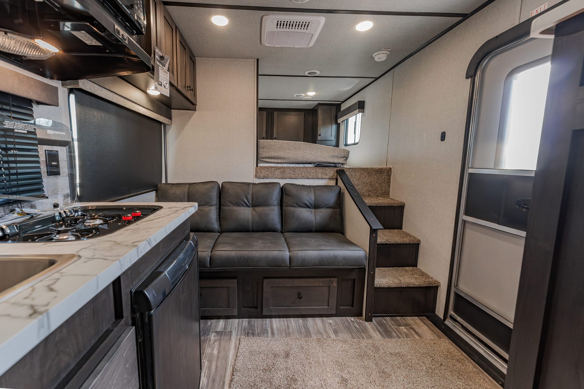 Interior of a compact RV: kitchen with stove on left, sofa in foreground, loft bed up stairs at rear, neutral tones and cabinets throughout
