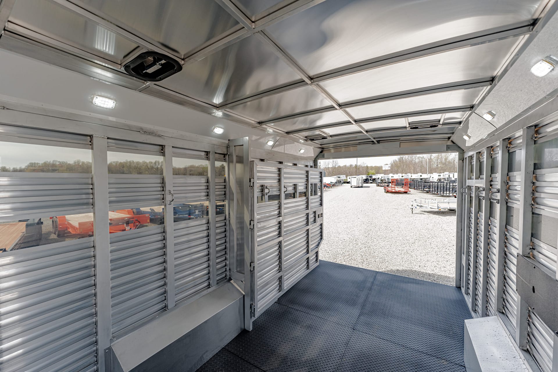 Interior of a silver metal trailer with ribbed walls, diamond-plate floor, and an open rear view showing a trailer yard outside.