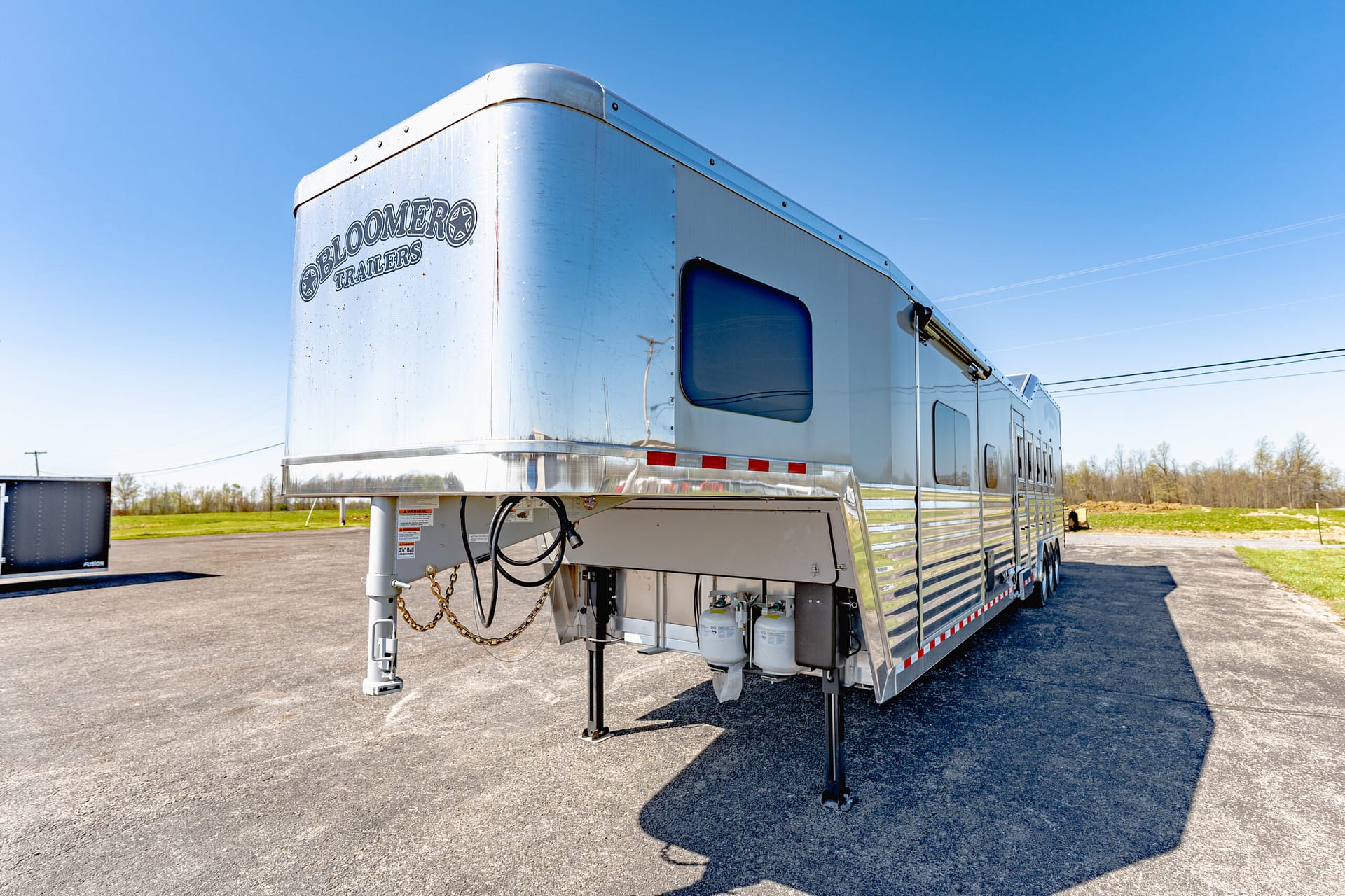 Shiny aluminum trailer viewed from the front-left, with 'Bloomer Trailers' logo, parked on a paved lot under a clear blue sky.