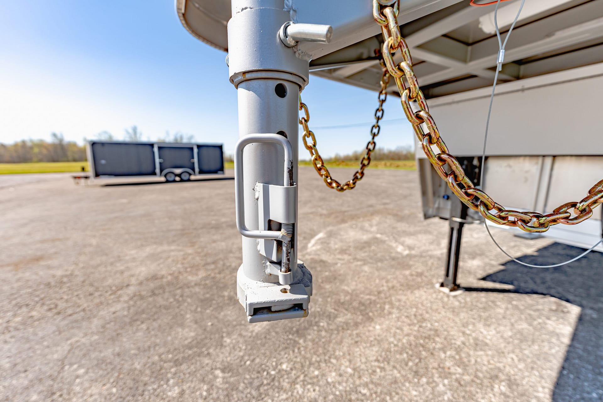Close-up of a gray trailer hitch post with a securing chain and hardware, a parked trailer visible in the background.