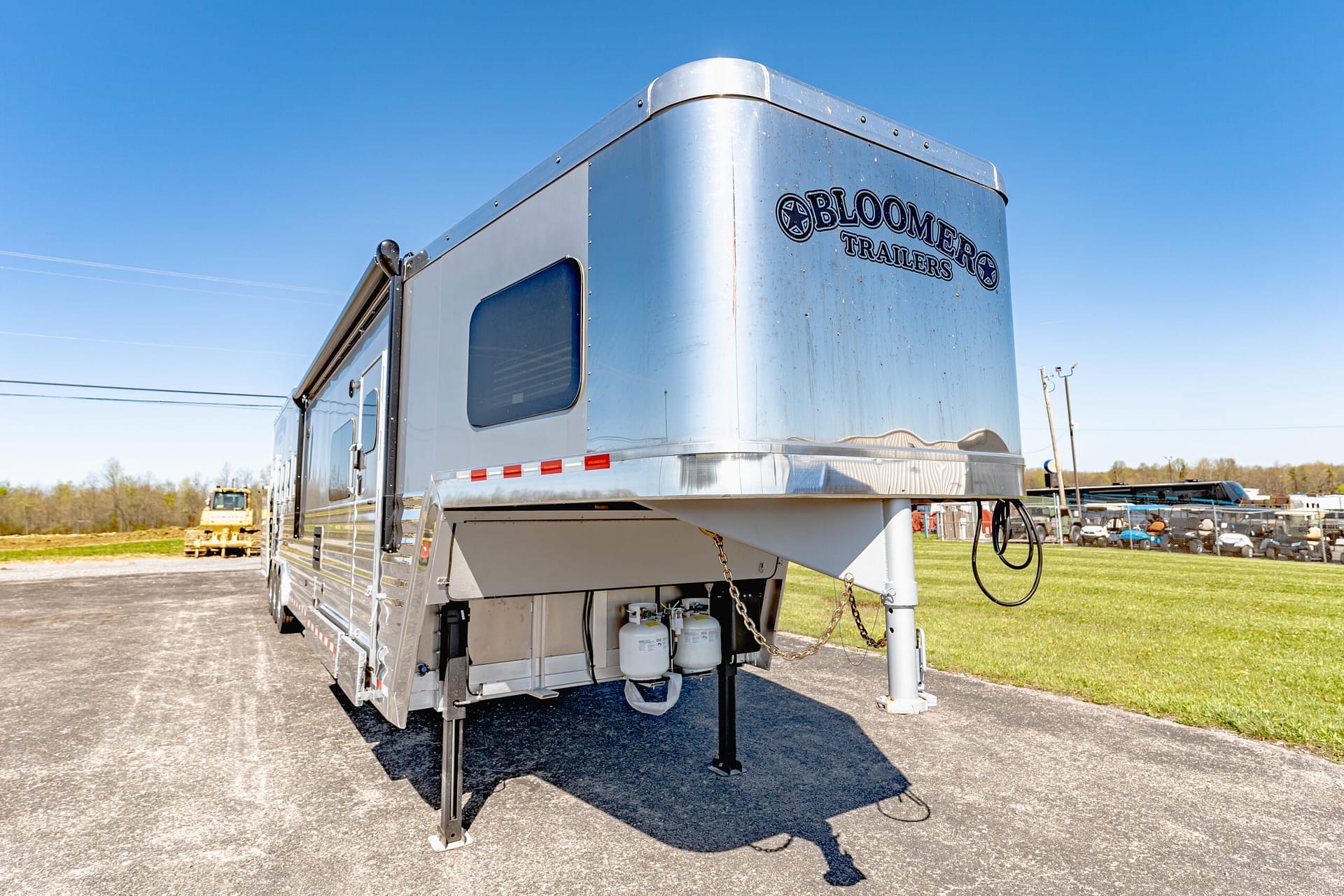 Side view of a shiny silver trailer with the Bloomers Trailers logo, parked on a lot under a clear blue sky; hitch and propane tanks visible underneath.