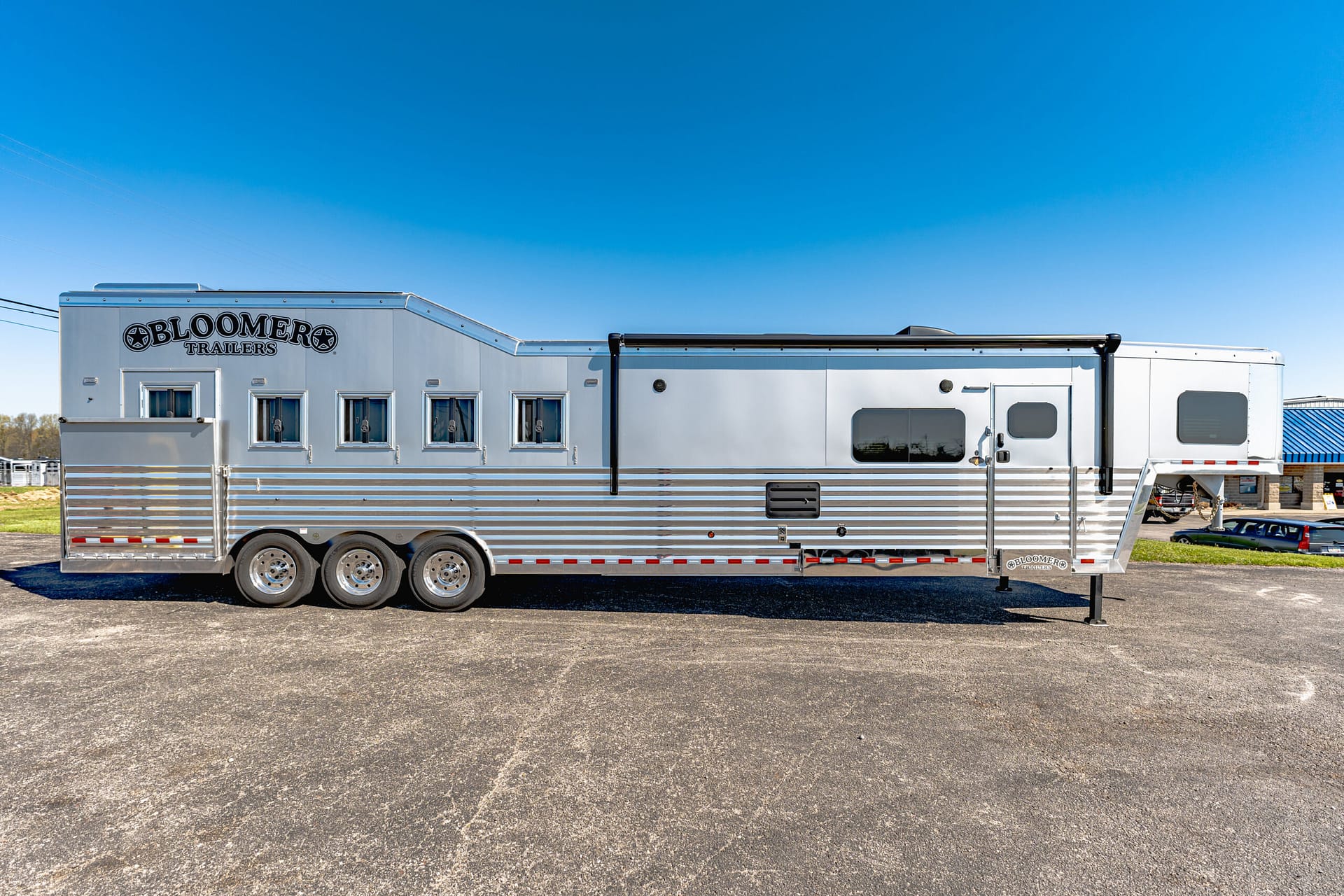 Silver Bloomer Trailers horse trailer parked on a paved lot under a clear blue sky.