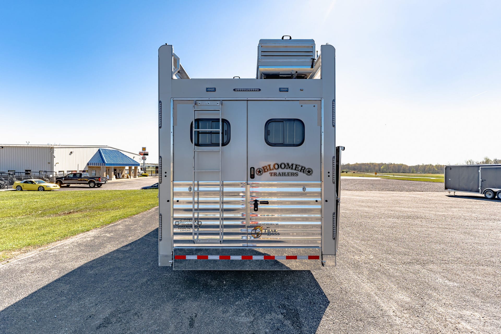 Rear of a large silver trailer with a side ladder, two rounded windows, and a blue-sky backdrop in a parking lot.