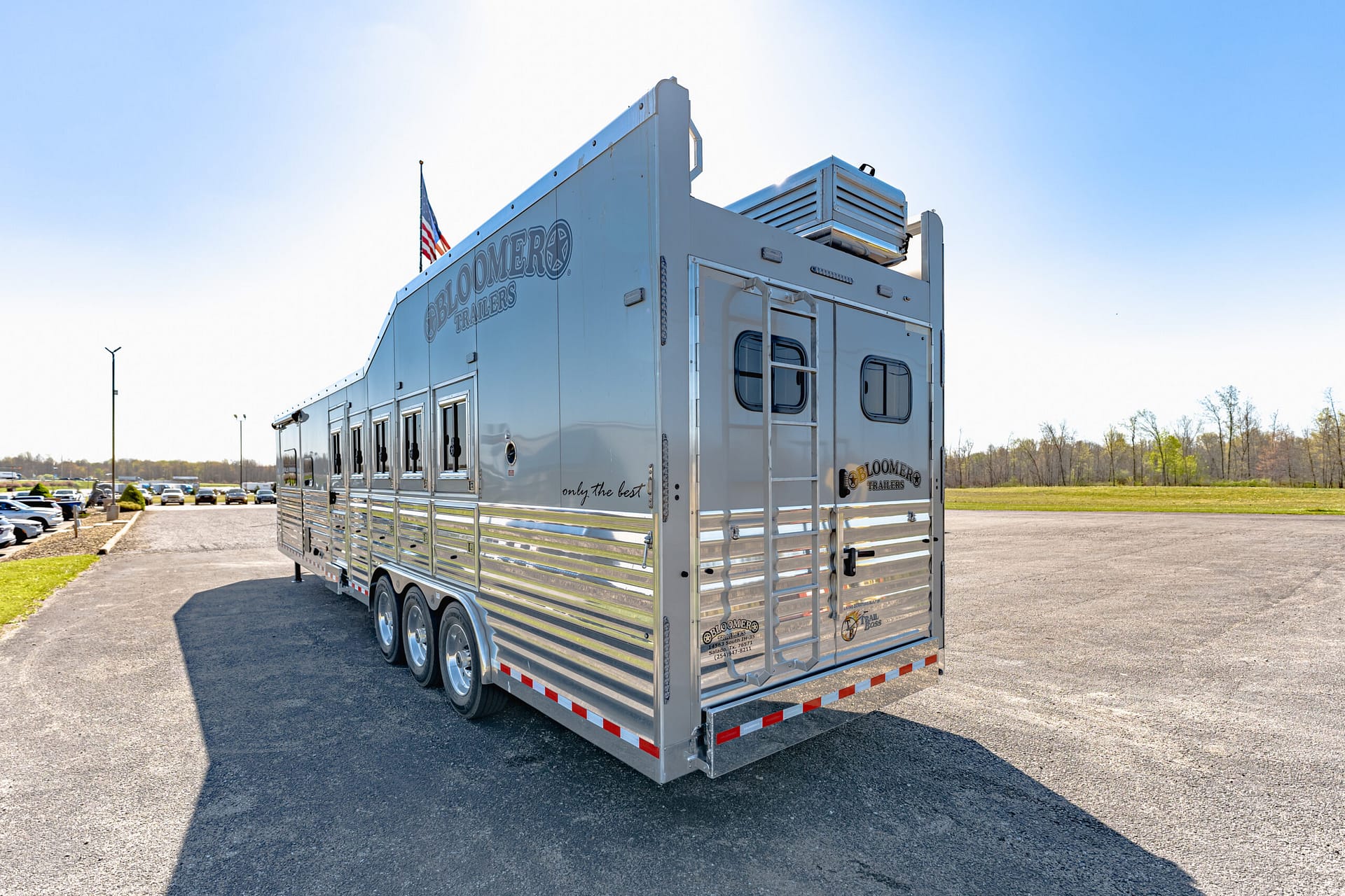 Silver livestock trailer parked in a lot under a clear blue sky, American flag flying atop and row of vehicles in the background.