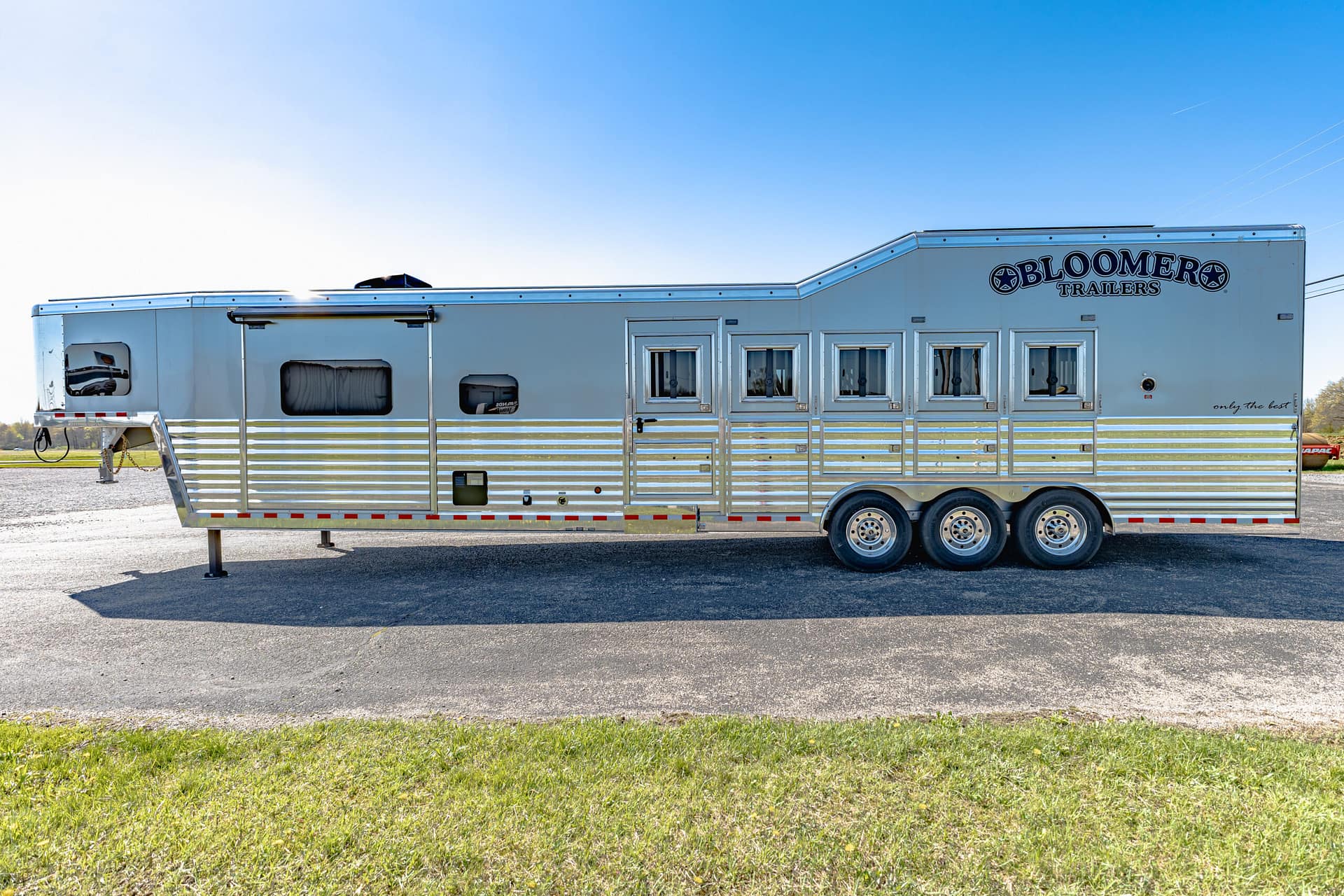 Silver Bloomer horse trailer with multiple windows parked on a paved lot under a blue sky.