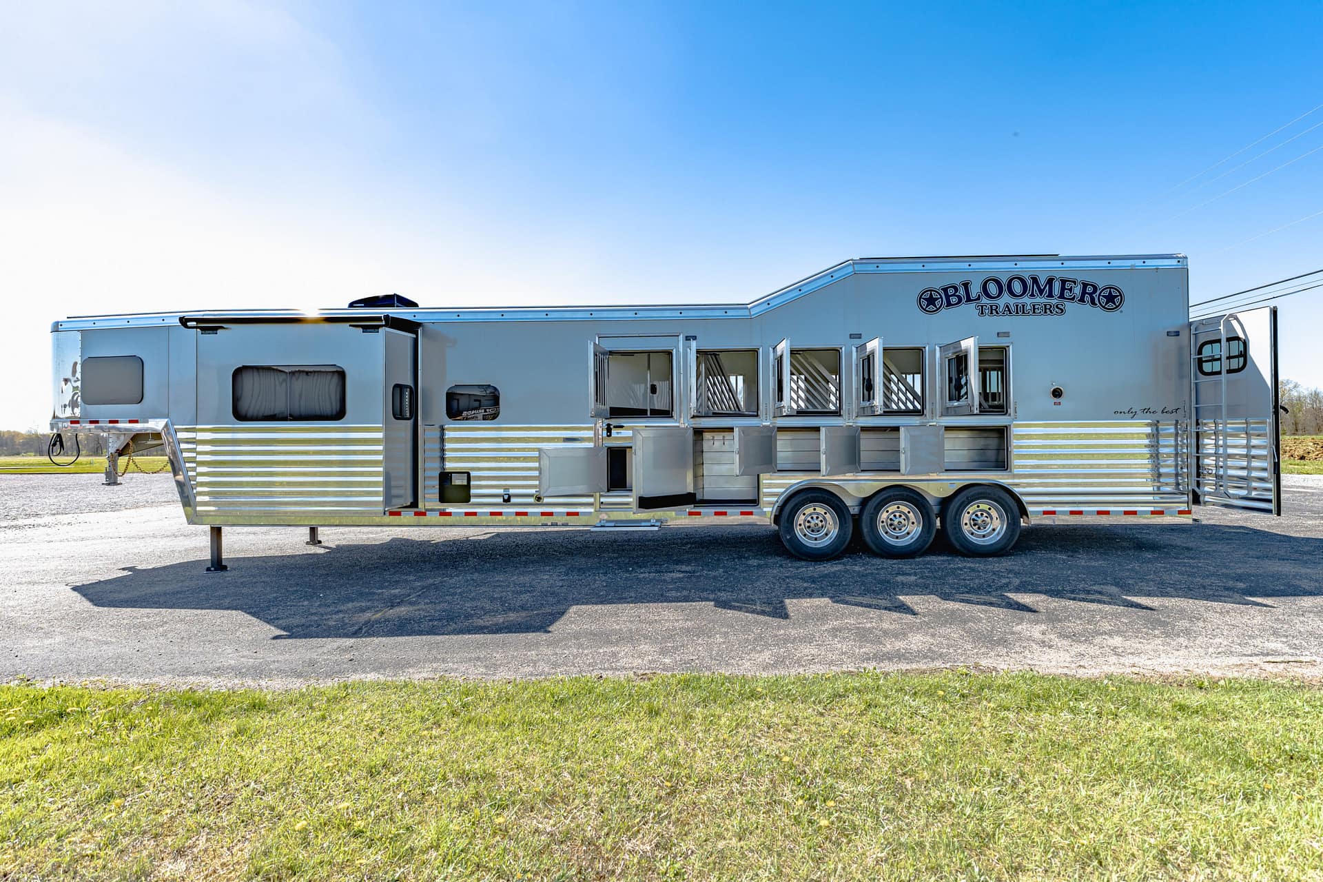 Long silver Bloomer Trailers recreational trailer on a gravel lot under a clear blue sky, with several windows and compartments visible on the side.