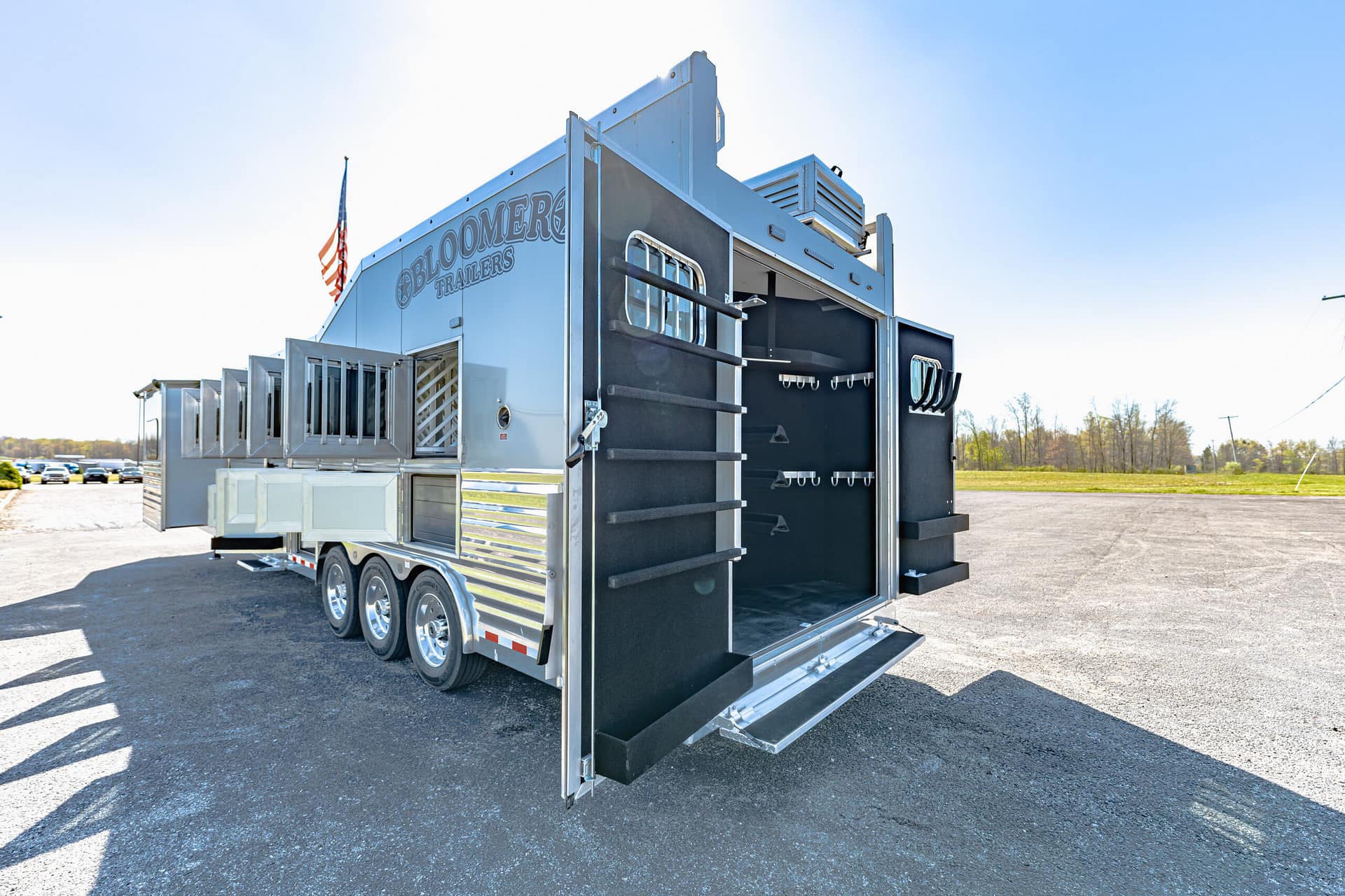 Large silver trailer with open rear doors exposing interior racks and hooks, parked on an empty lot under a blue sky