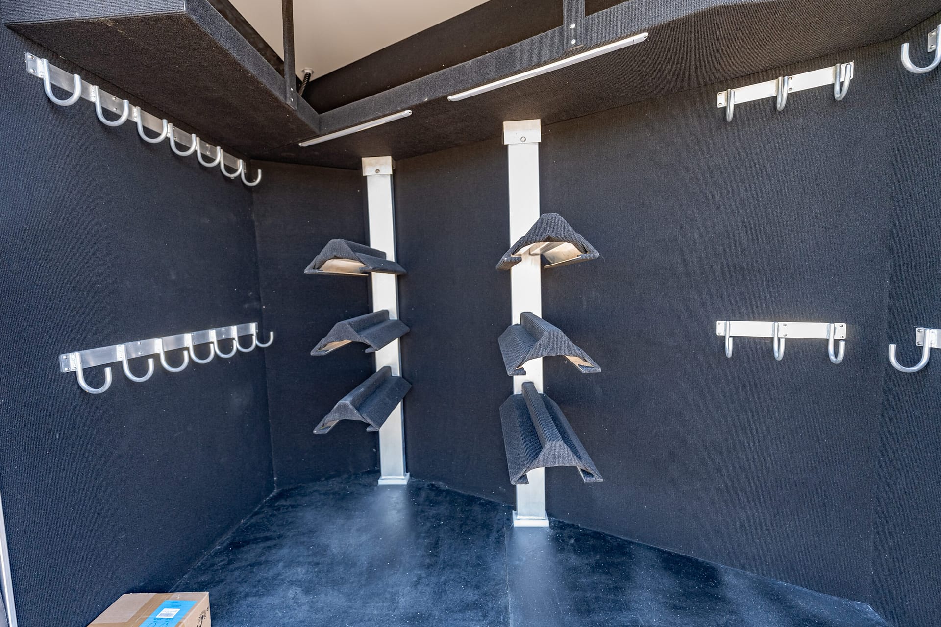 Interior of a dark cargo trailer with numerous silver U-shaped hooks along the walls, vertical metal supports, and a single cardboard box on the floor left corner.
