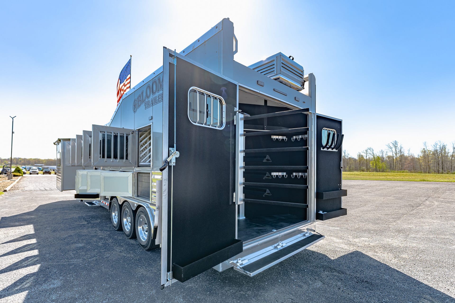 Large silver and black trailer with an open rear ramp and gear racks, American flag flying on a sunny day in a parking lot.