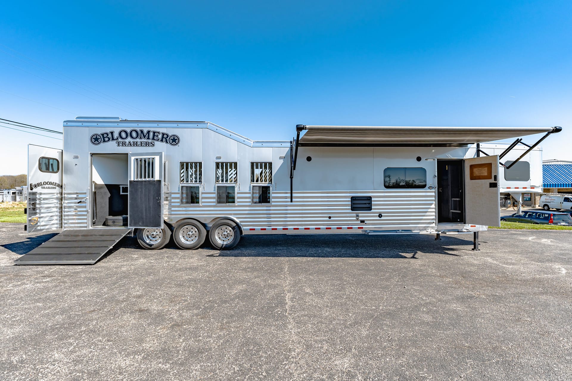 White Bloomer Trailers horse trailer with ramp down in a parking lot and a clear blue sky above.