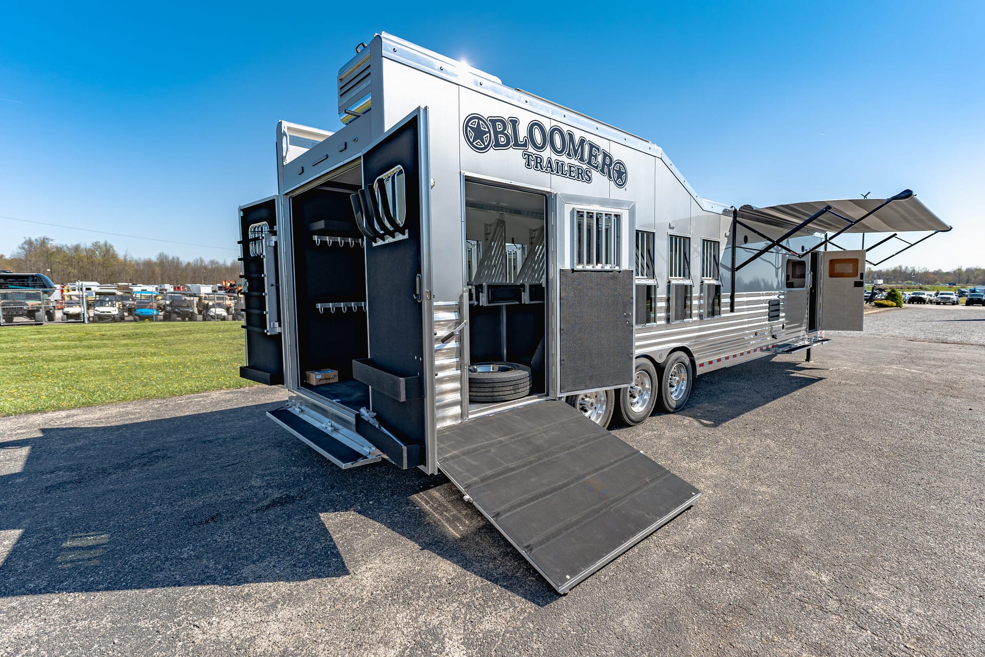 Shiny silver Bloomer Trailers vehicle with multiple storage doors open and a large ramp extended in a parking lot area.