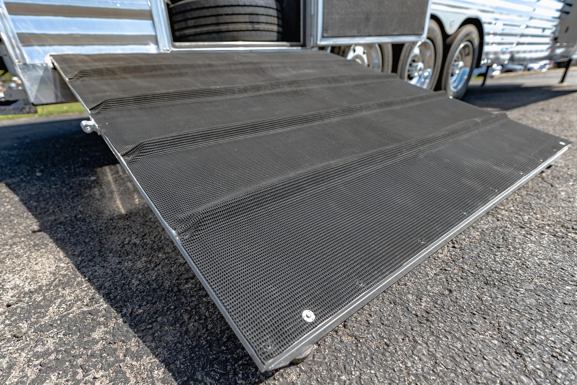 Metal loading ramp attached to a silver trailer, featuring a black textured non-slip surface and visible trailer wheels in the background beneath a sunny sky.
