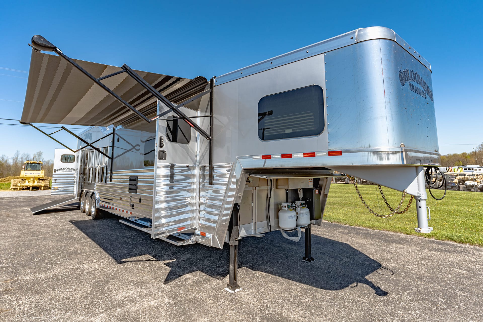 Silver travel trailer with an extended striped fabric awning, parked on a paved lot under a bright blue sky.