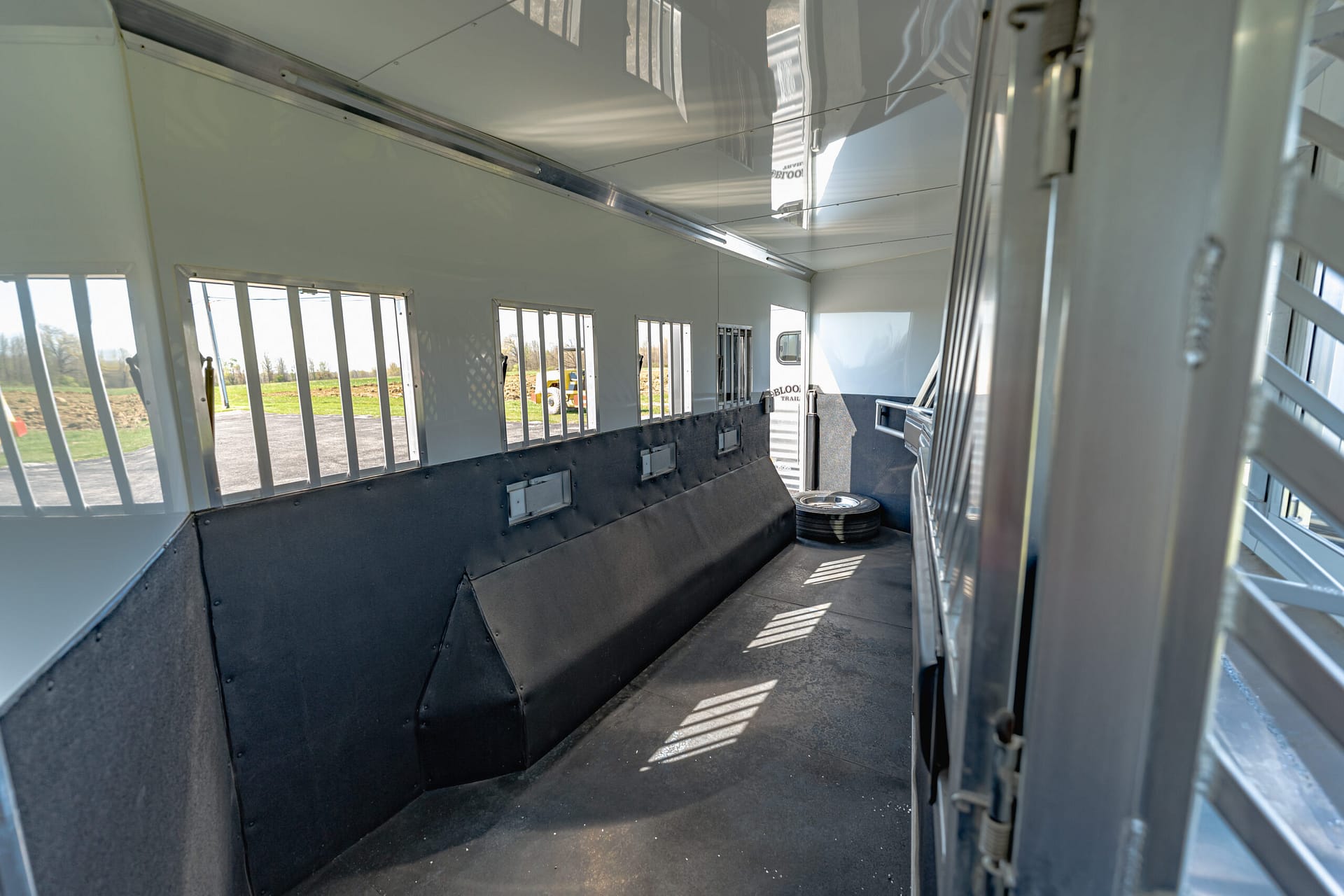 Interior of a white livestock/horse trailer with side windows, dark padded lower walls, and a spare tire on the floor near the back.