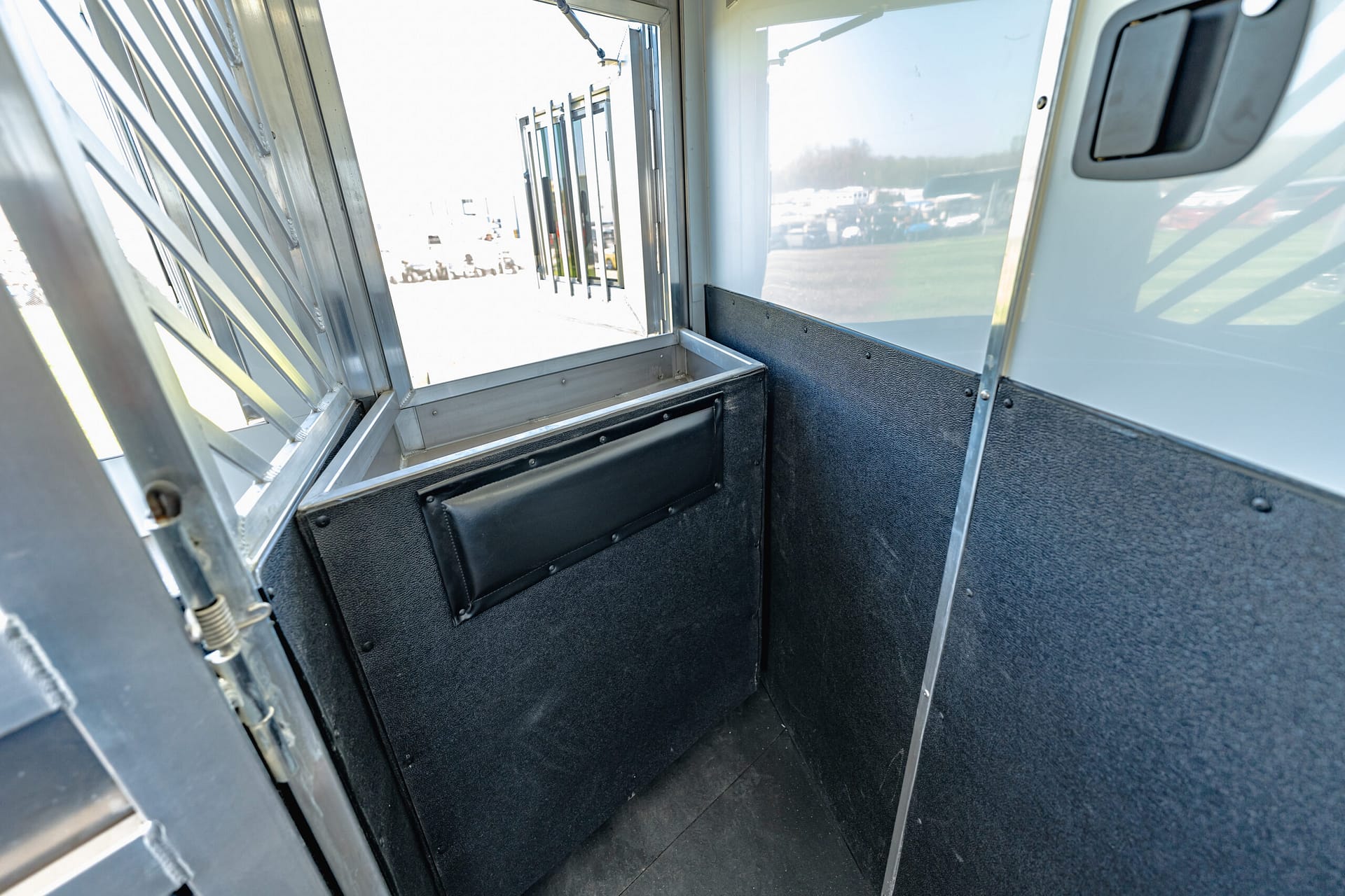 Interior view of a small metal service booth with a black padded bench and metal walls; open window with vertical bars, parking lot visible outside