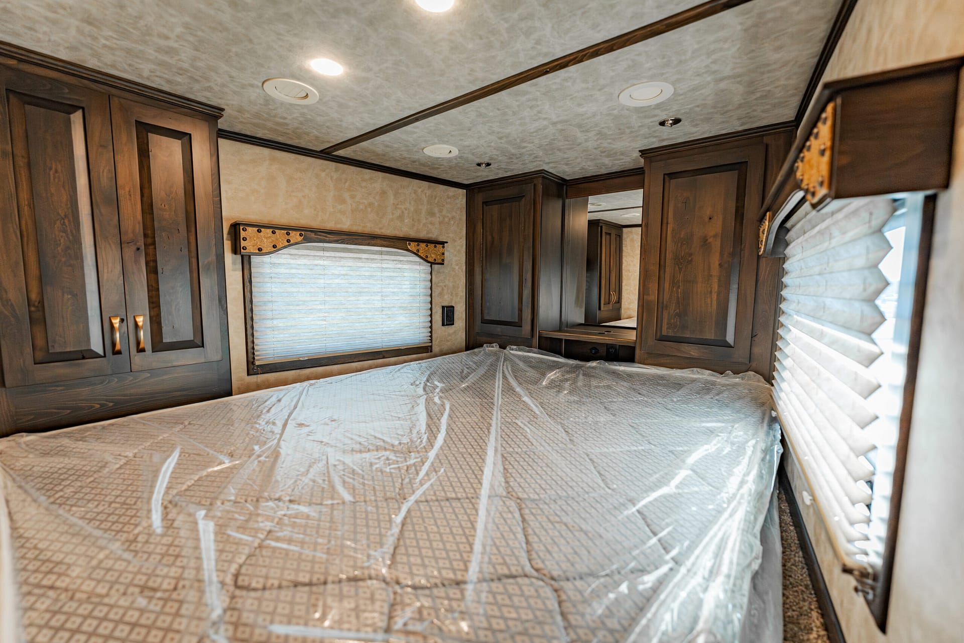 Interior of an RV bedroom with dark wood cabinets, a window with blinds, and a bed covered in protective plastic.