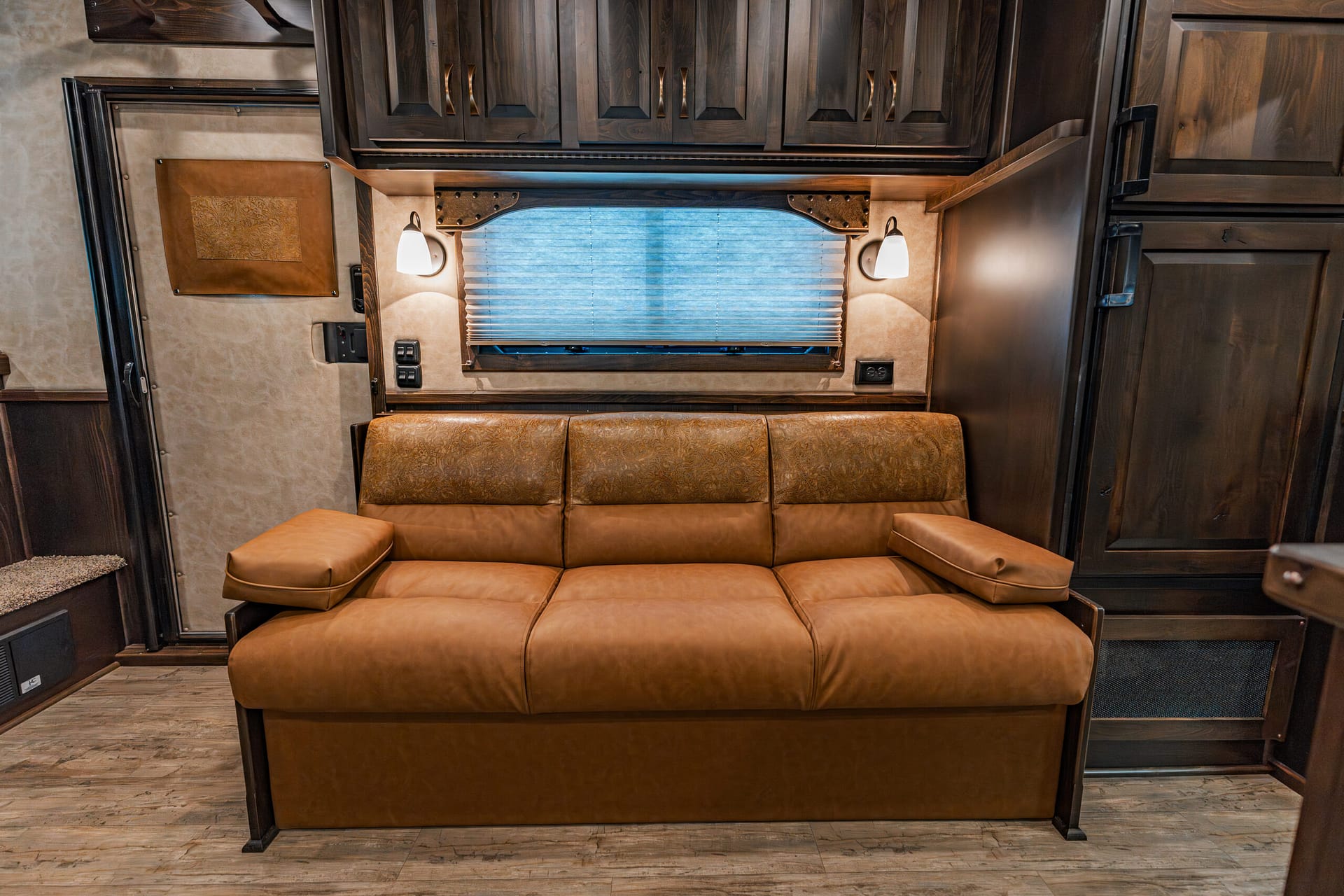 RV living area with tan leather sofa, dark wood cabinets above, and a window with pleated blinds flanked by two wall lamps.