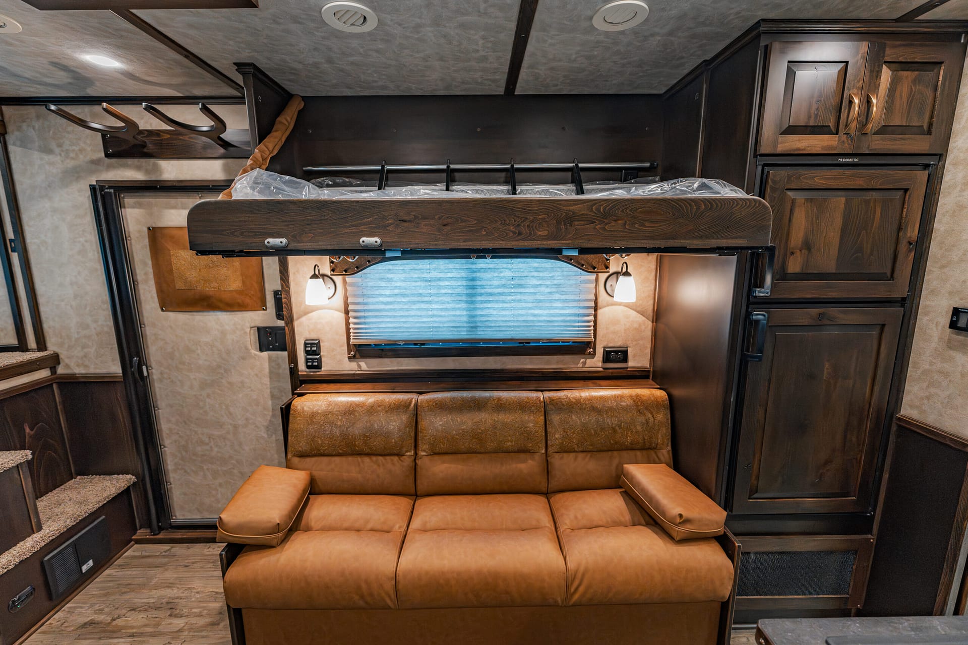 RV interior with a top bunk bed above a brown leather sofa and overhead wood cabinets in a compact living area