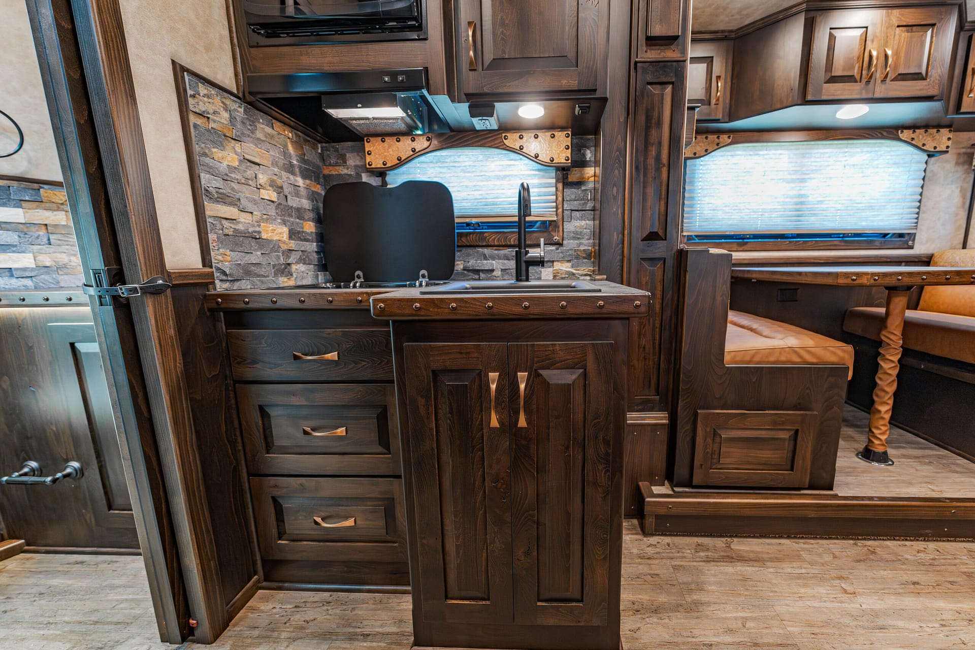Interior of a RV kitchen with dark wood cabinets, stone backsplash, and a black sink area under a window with pleated blinds.