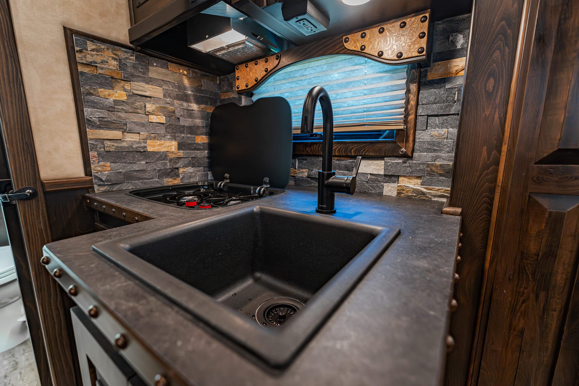 Compact RV kitchen: black sink and faucet, two-burner stove, stone tile backsplash, and decorative wood panels.”] ,