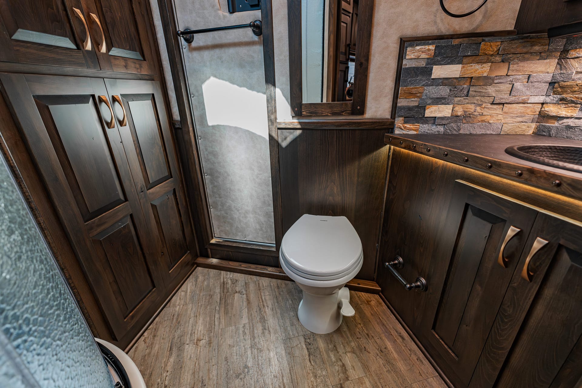 Tiny RV bathroom with dark wood cabinetry, a white toilet, and a stone-tile backsplash while a mirror frames the vanity area