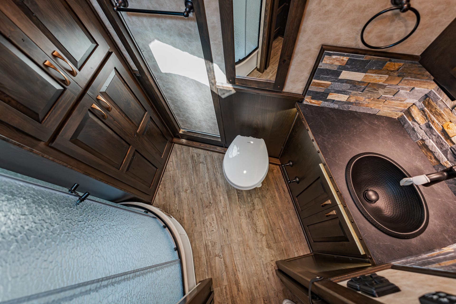 Compact RV bathroom with dark wood cabinets, a white toilet, and a frosted glass shower on the left; stone-tile accent wall on the right.
