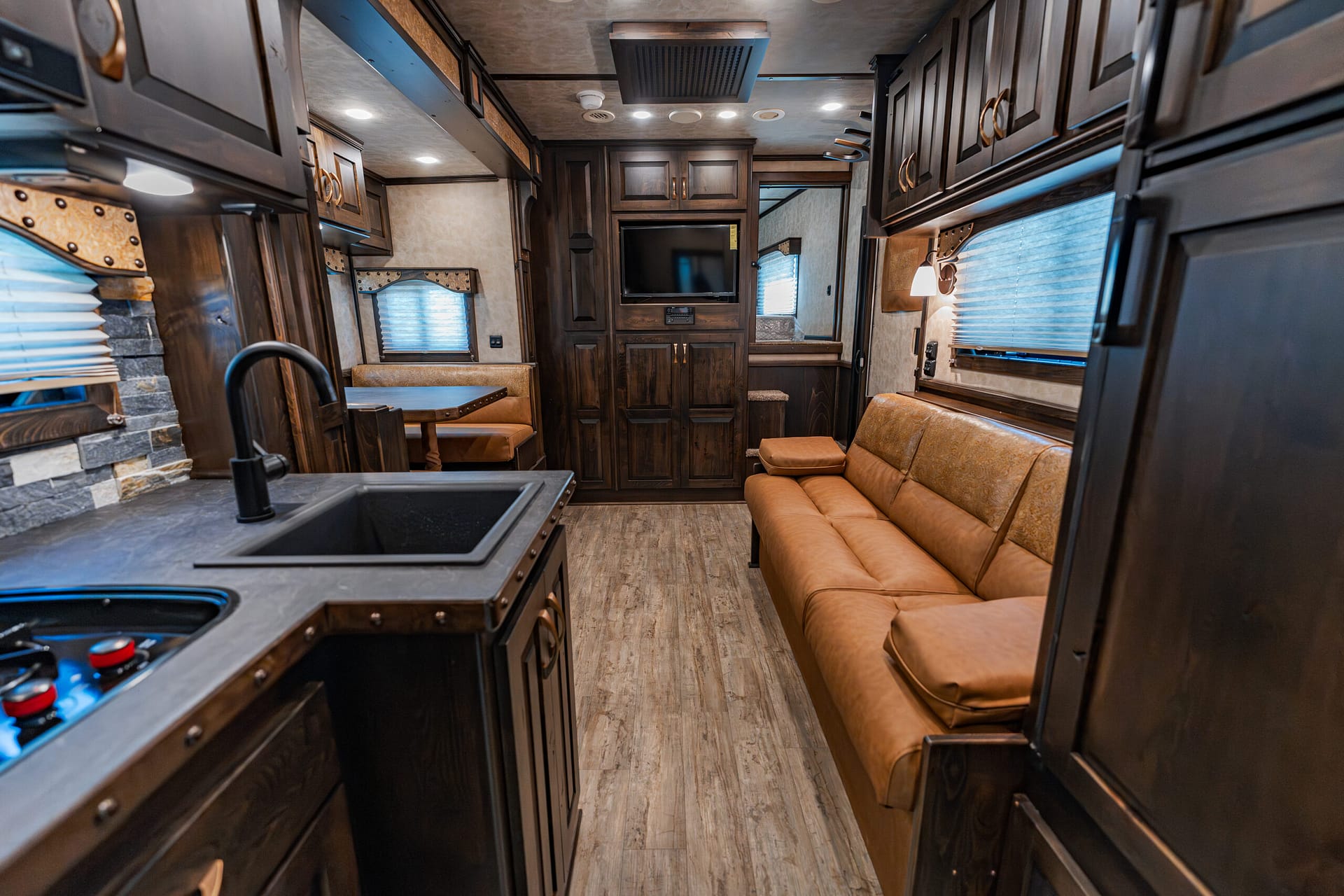 Interior of a modern RV living area with dark wood cabinets, tan leather sofa, and a small dining table by the window while the kitchen is on the left.