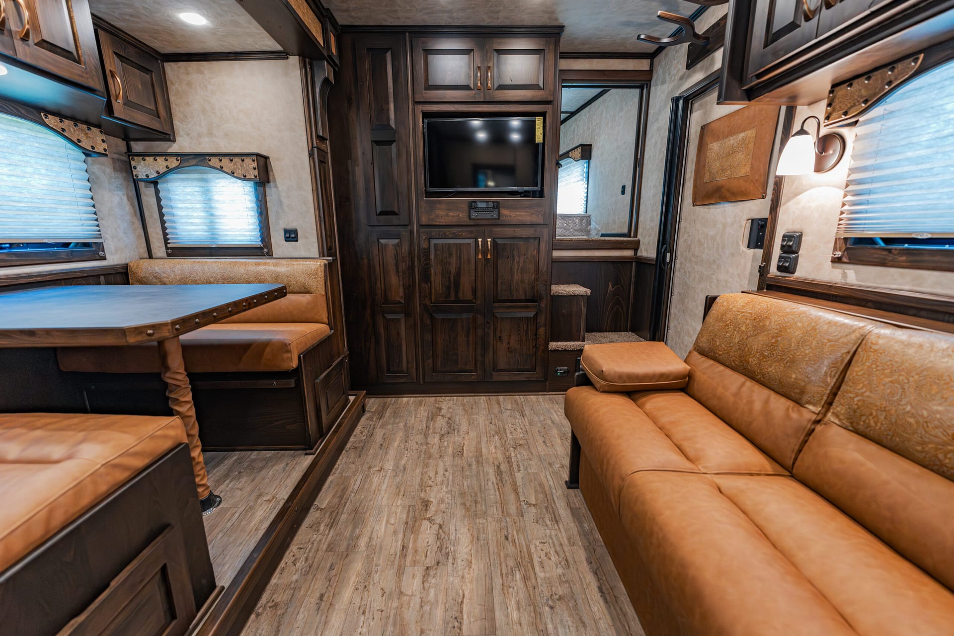 Cozy RV living area with dark wood cabinets, tan leather seating, a central entertainment wall with a TV, and windows with blinds.