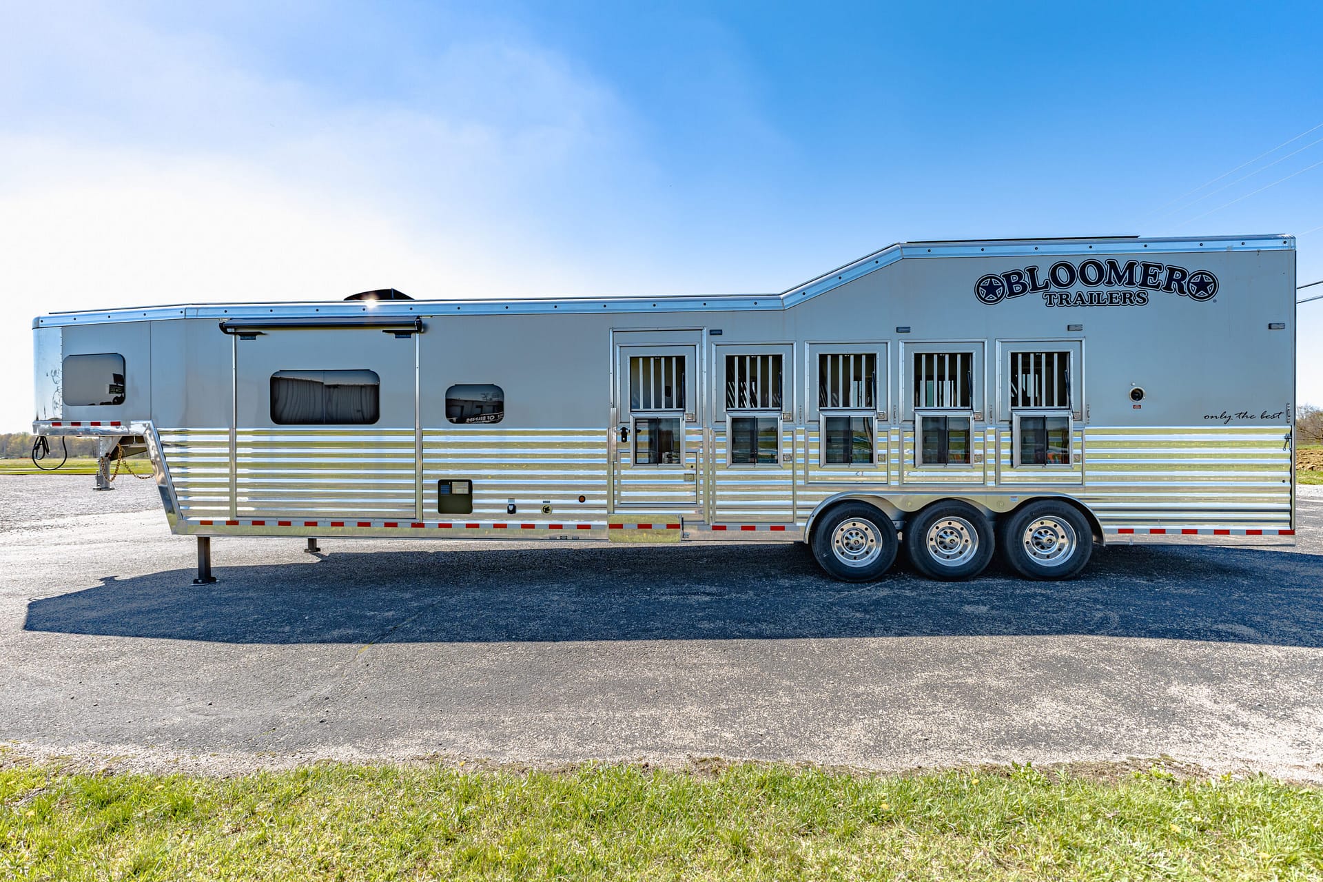 Side view of a long silver Bloomer Trailers horse trailer parked on a gravel lot under a clear blue sky, with multiple windows and two axles visible.