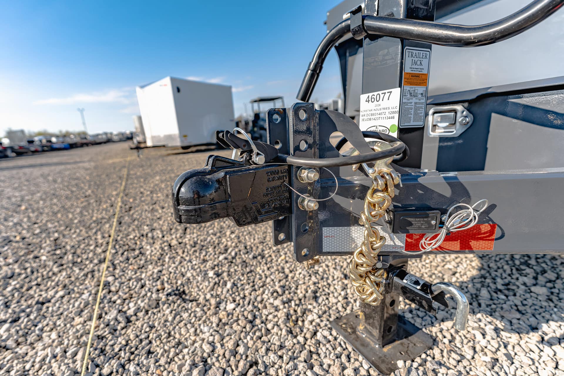 Close-up of a trailer hitch with safety chains and wiring on a gravel lot, with other trailers blurred in the background.
