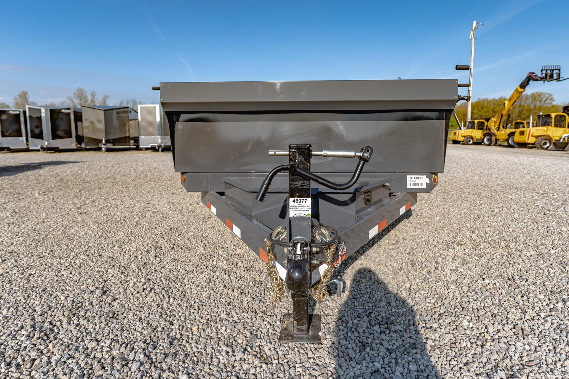 Front view of a gray dump trailer on a gravel lot, showing the hitch and safety chains with a clear blue sky above.