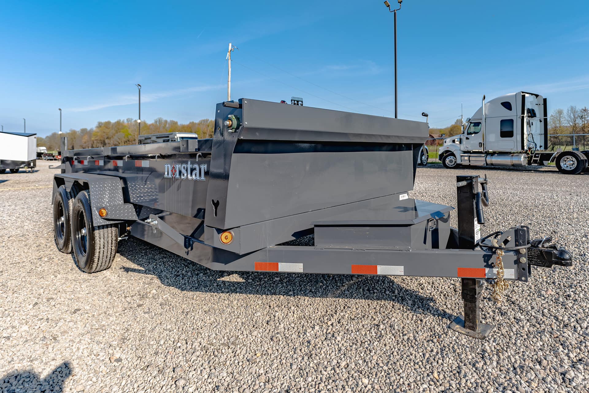 Gray heavy-duty trailer with dual axles in a gravel lot, blue sky overhead, and a white semi-truck in the background