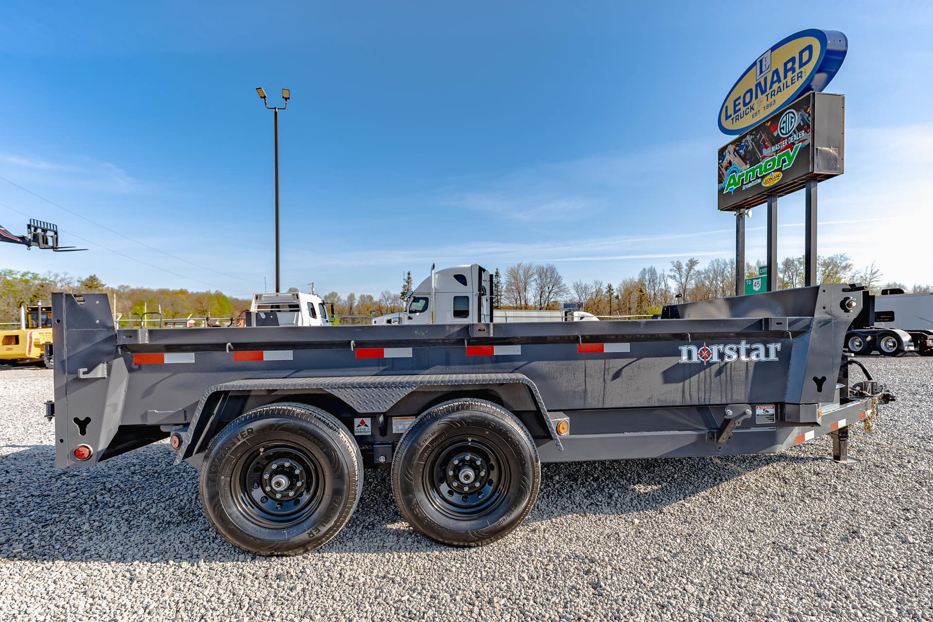 Gray multi-axle trailer on a gravel lot with a bright blue sky and a tall Leonard trailer sign in the background, Norstar branding on the side.