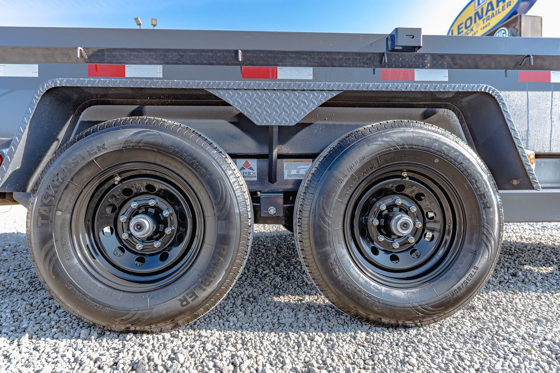 Close-up of dual trailer wheels with black steel rims and a diamond-plate fender, on a gravel surface.