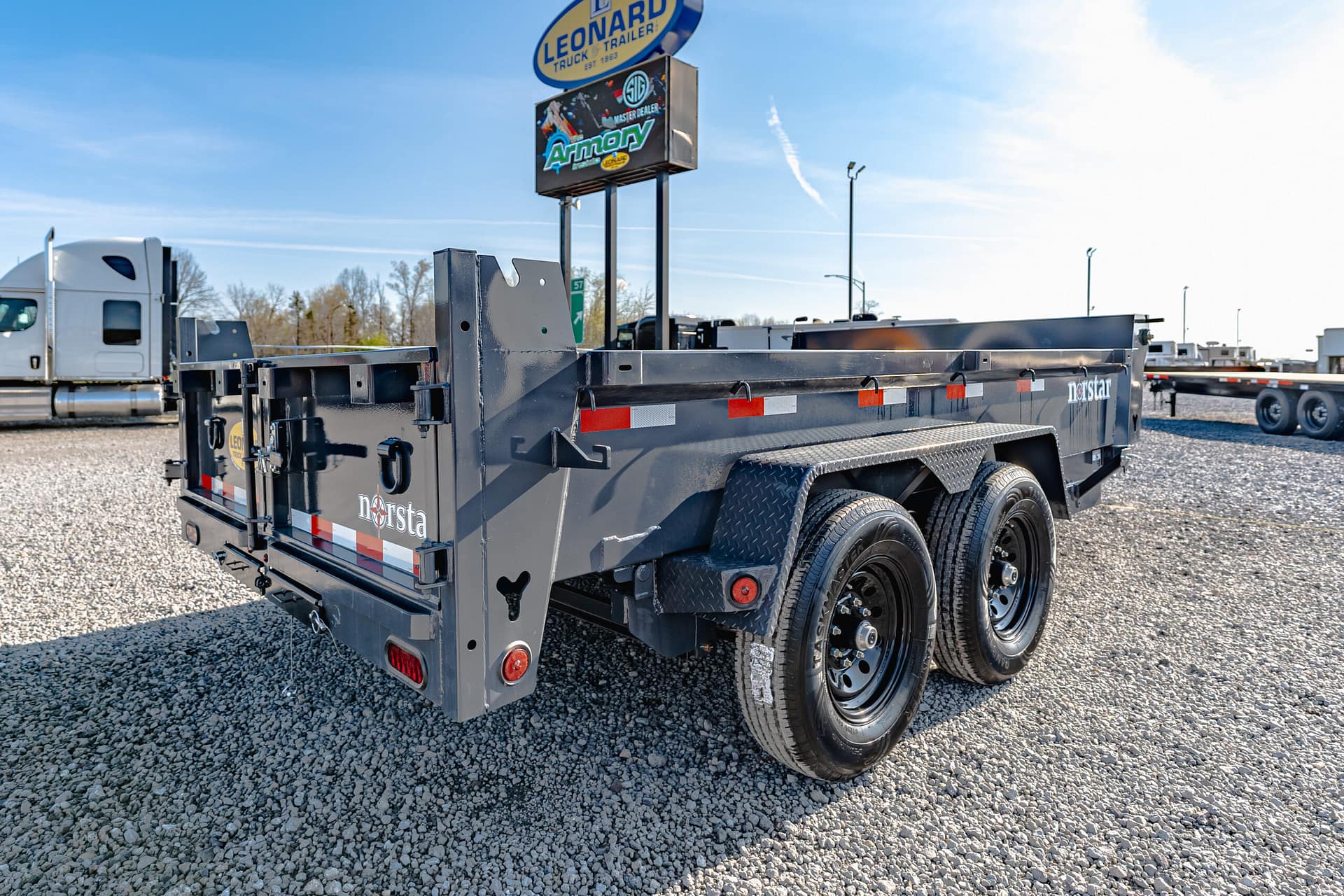 Black steel flatbed trailer with dual rear axles in a gravel truck yard under a clear blue sky. The scene shows an industrial trailer with reflective safety stripes and equipment mounts at the rear.