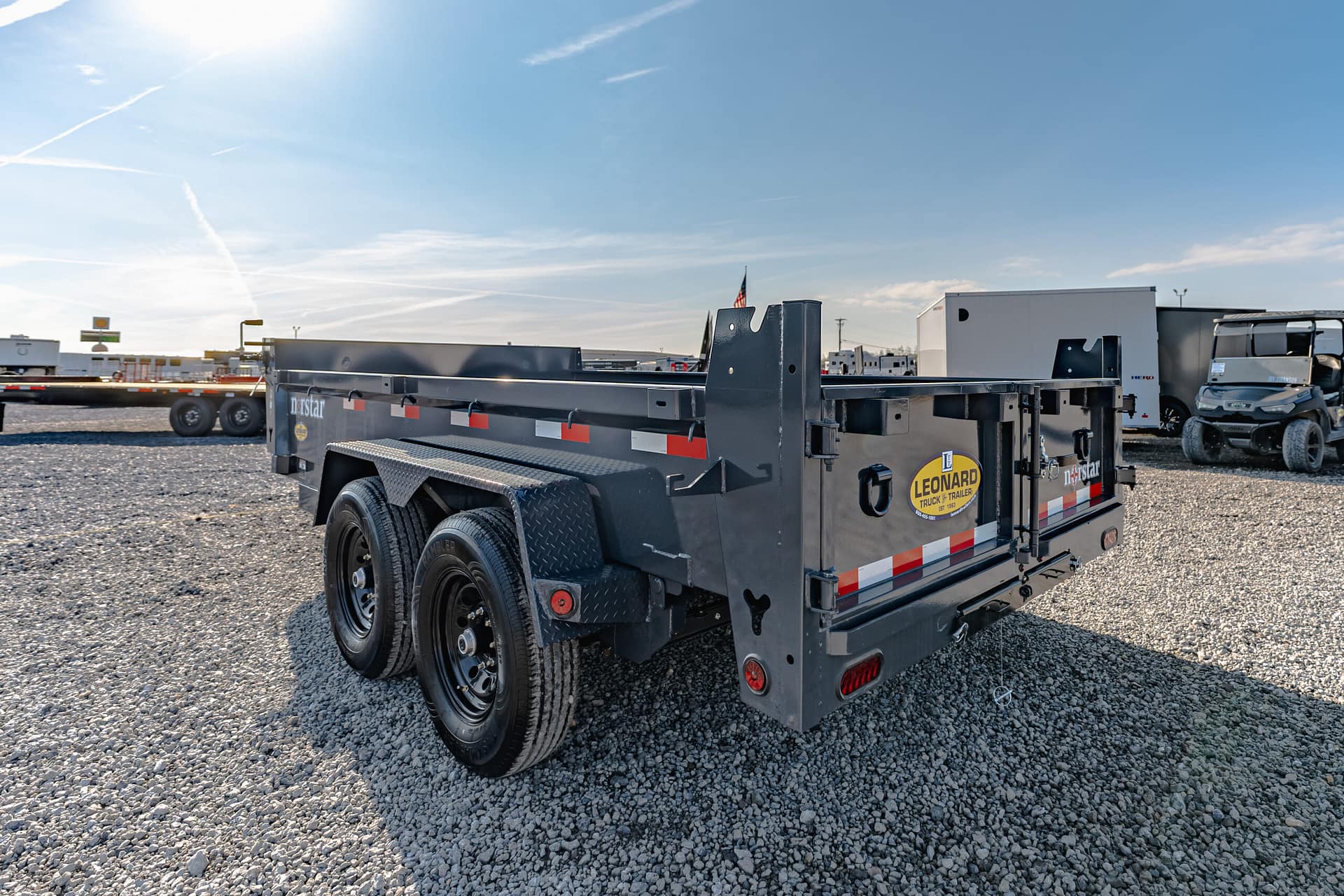 Grey flatbed trailer in a gravel lot with an American flag and other trailers in the background, bright sunny day.
