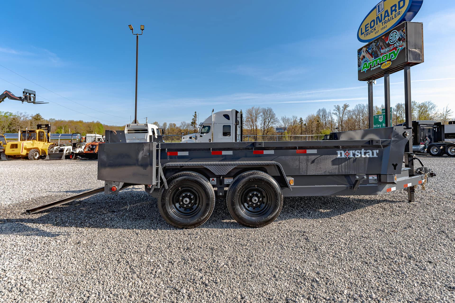 Gray flatbed trailer with dual axles on a gravel lot, with rail tracks in the foreground and industrial equipment in the background beneath a clear blue sky.