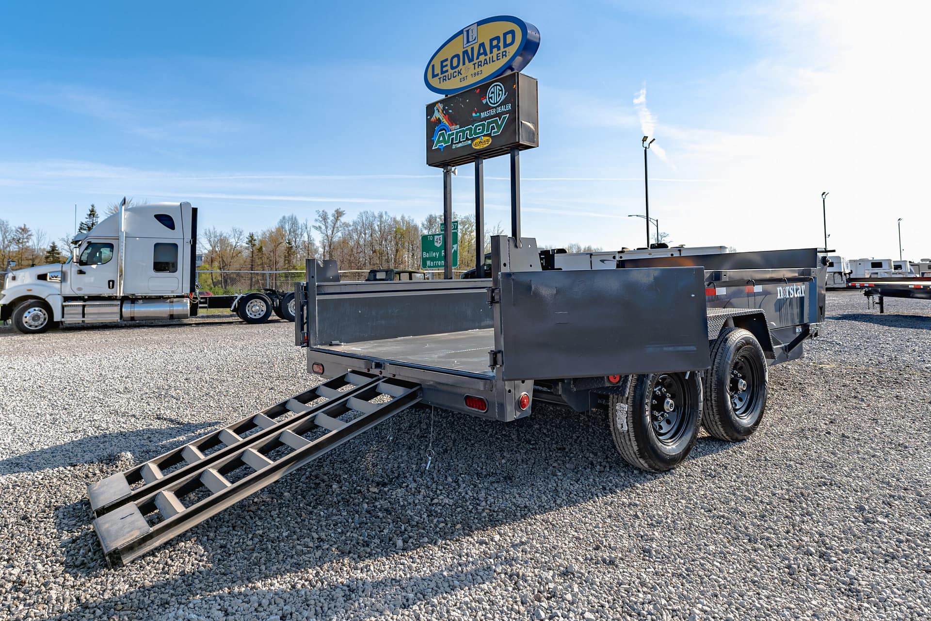 Flatbed trailer with metal loading ramps extended on a gravel lot, white semi-truck in the background under a clear blue sky