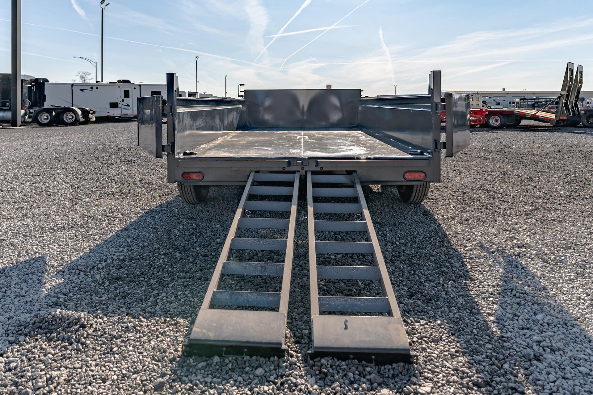 Rear view of a gray flatbed trailer with fold-down loading ramps, on a gravel lot with other trailers in the background under a blue sky.