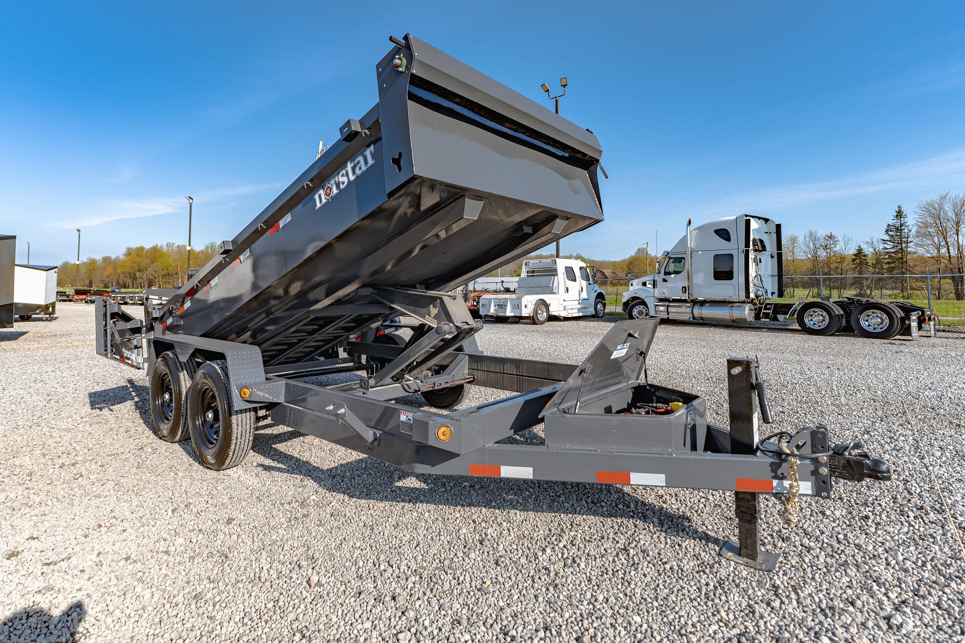 Gray dump trailer lifted at a truck yard, hydraulic bed angled for dumping, gravel ground and clear blue sky in the background.