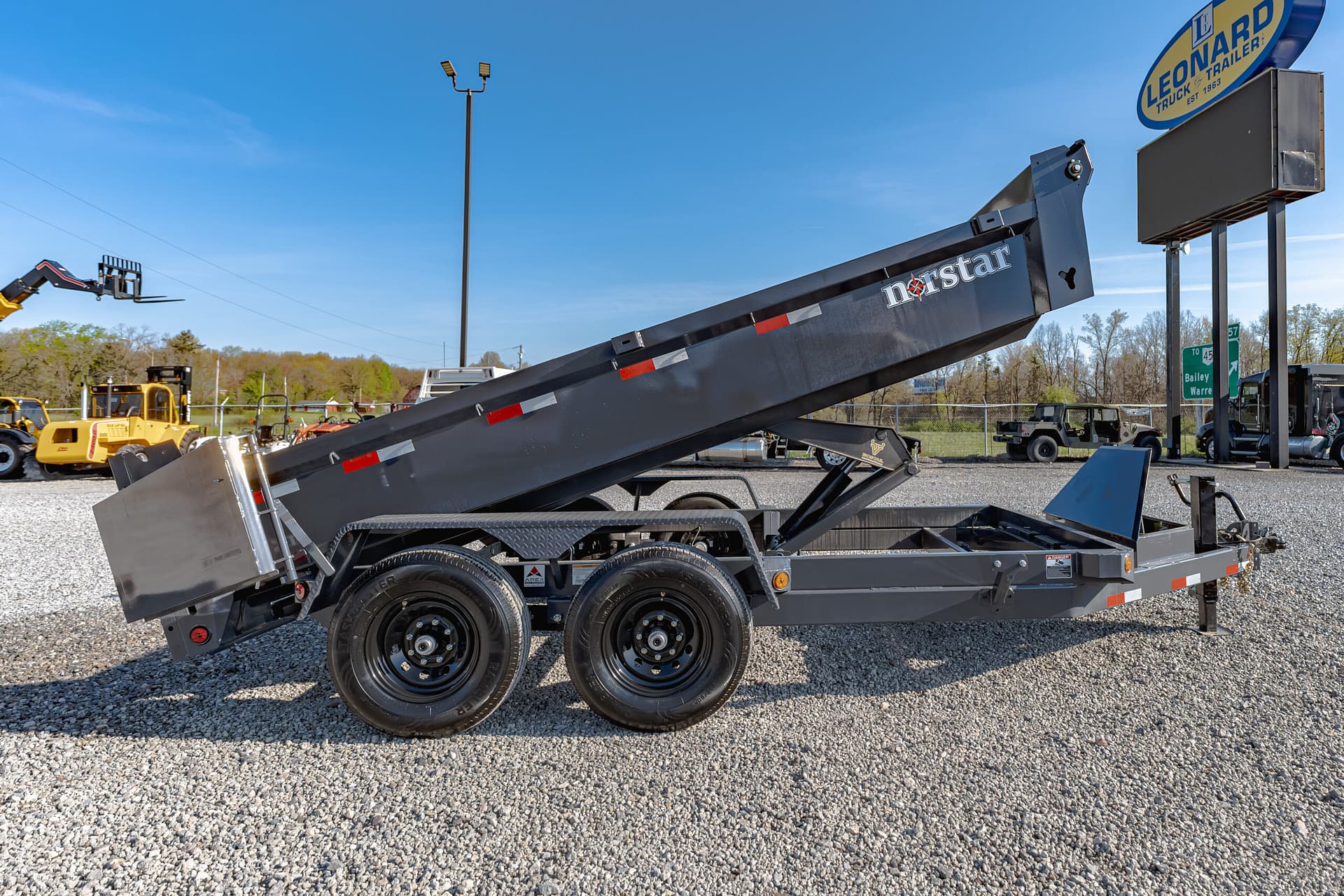 Gray Norstar tilt-bed trailer with raised bed and dual axles in a gravel lot, construction equipment in the background under a clear blue sky.
