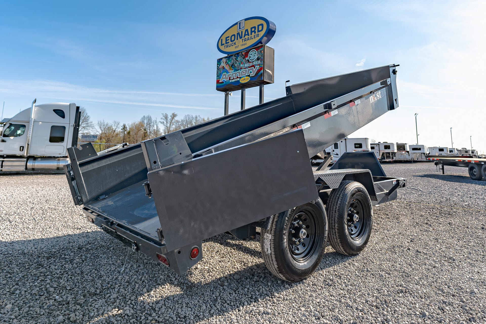 Tilted black steel dump trailer on a gravel lot with a white semi truck in the background and a Leonard sign overhead, under a clear blue sky.