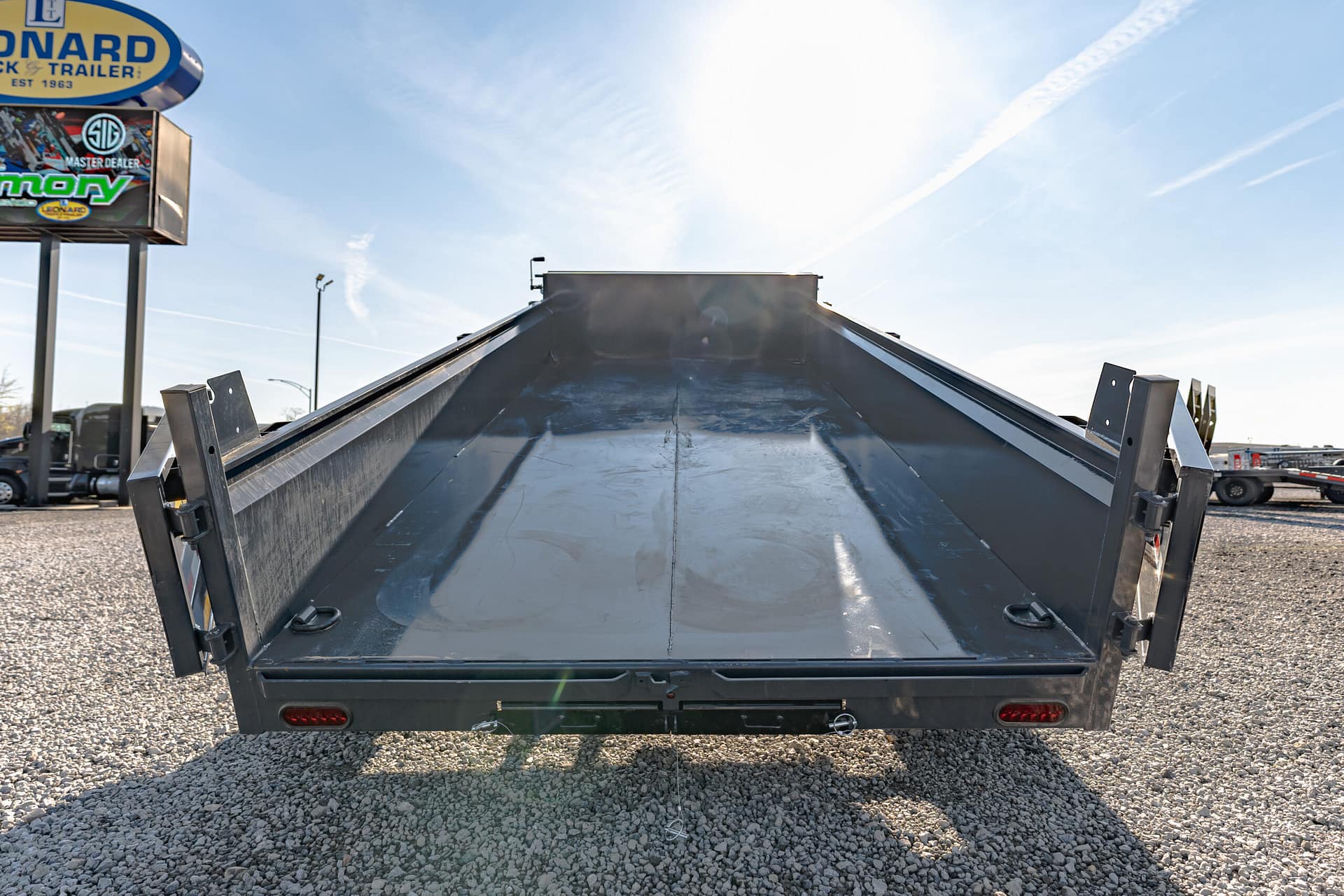 Rear view of a metal dump trailer with its bed raised, on a gravel lot; storefront sign visible on the left.