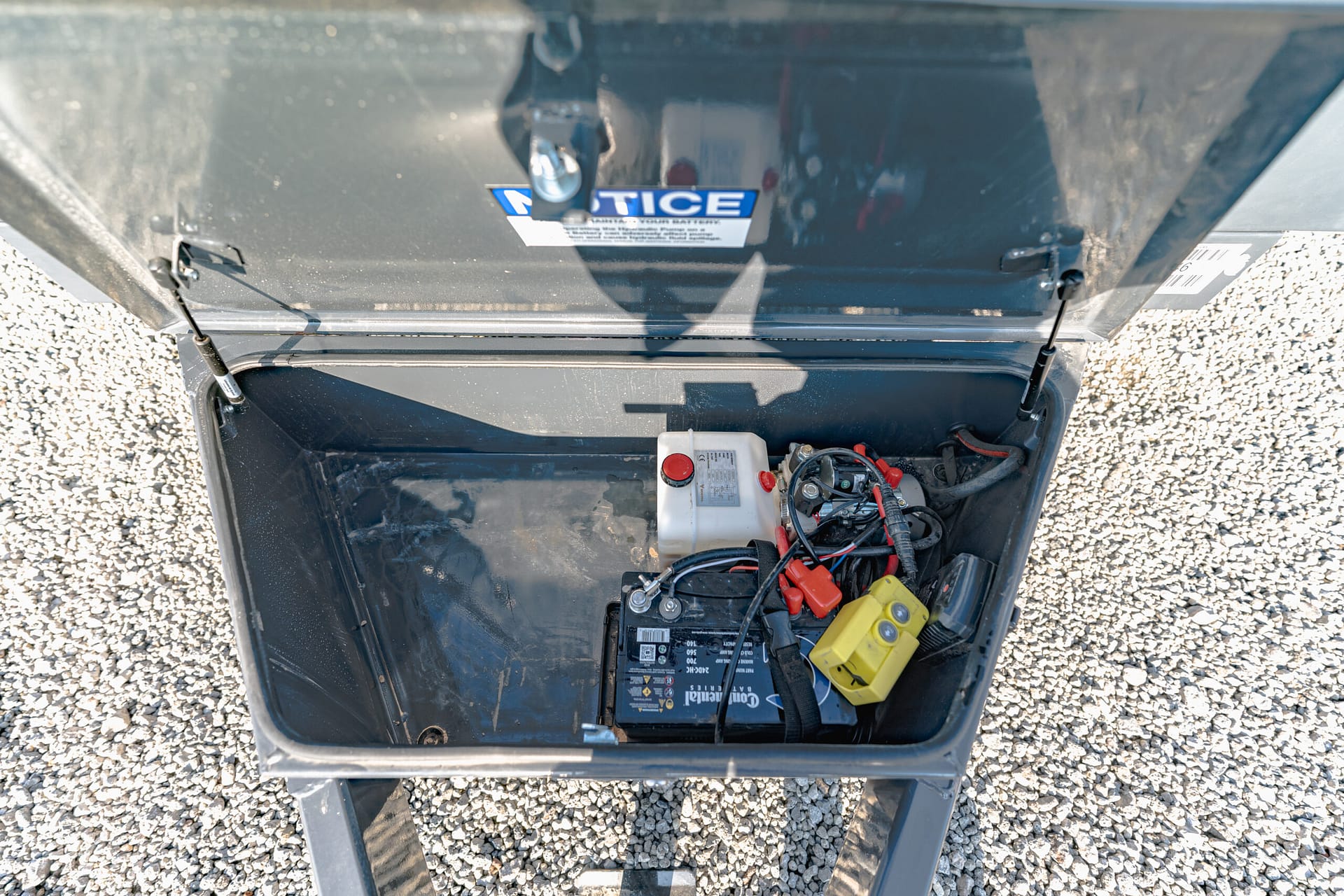 Open battery box containing a car battery with red and black cables, sitting on gravel surface.