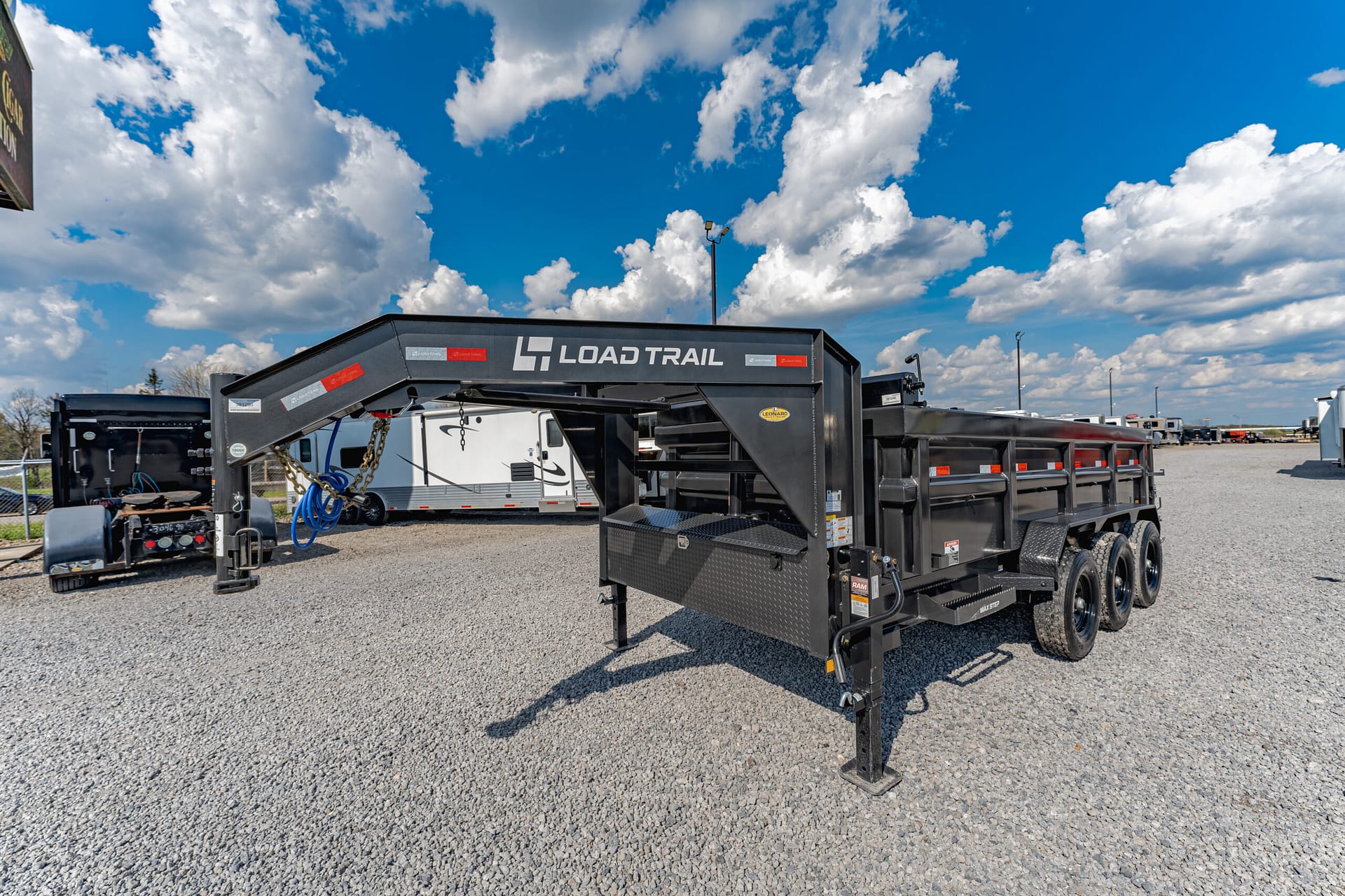 Black Load Trail flatbed trailer with dual axles parked on a gravel lot under a bright blue sky with scattered clouds; equipment and hoses visible nearby, suggesting a storage yard.