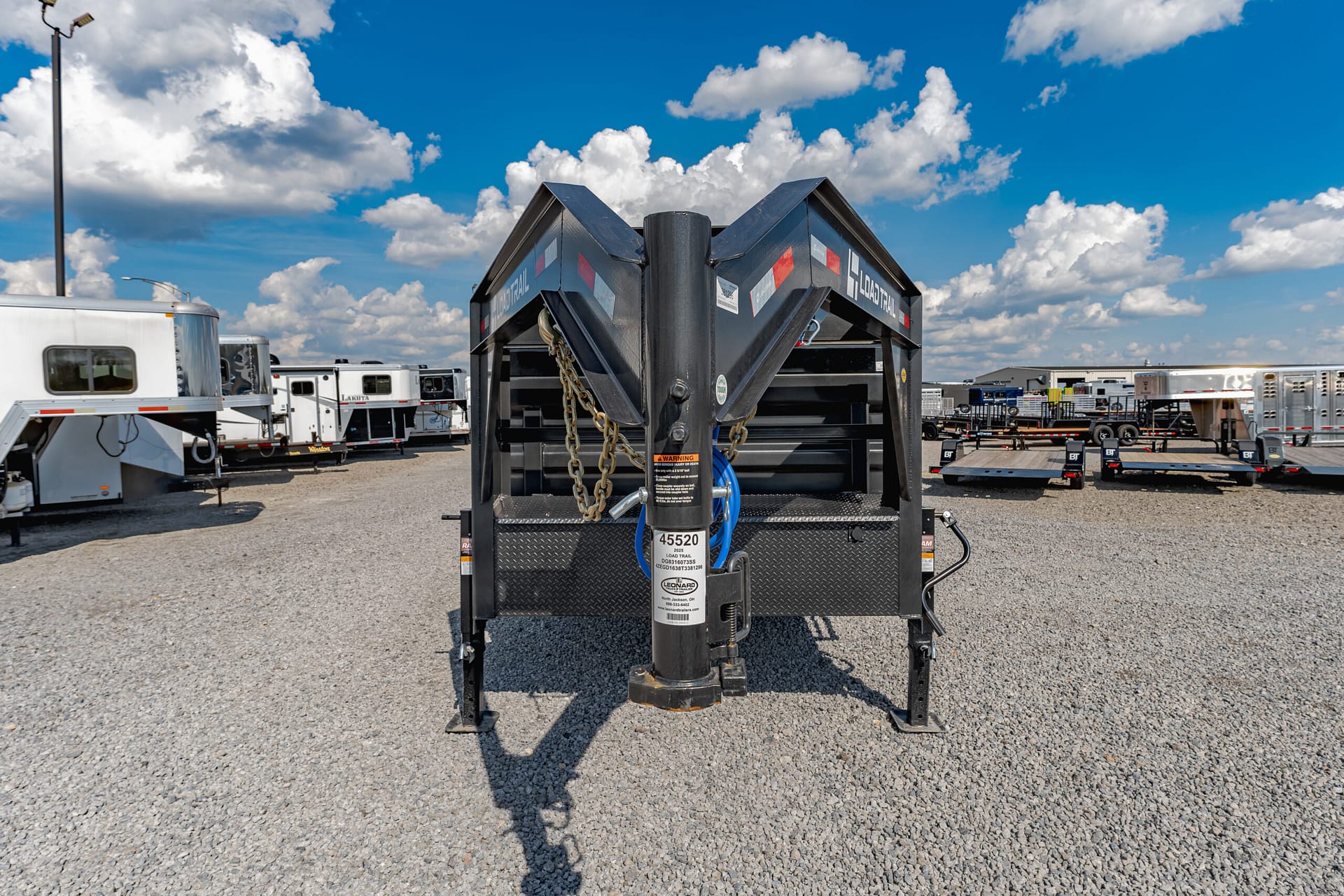 Close-up of a black gooseneck trailer hitch with safety chains in a gravel lot, surrounded by parked trailers.