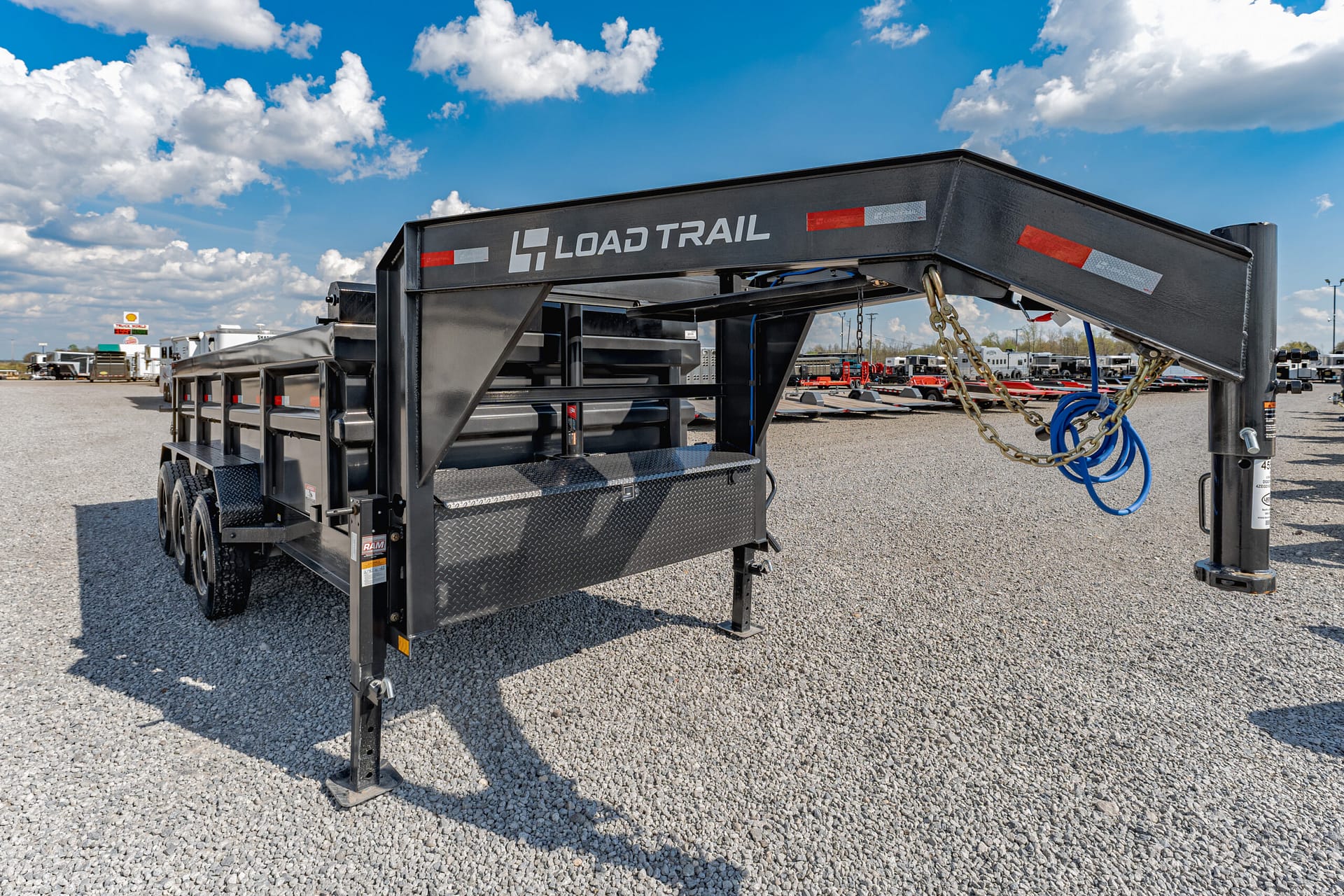 Black Load Trail gooseneck trailer with safety chains and blue hoses in a gravel lot under a blue sky.