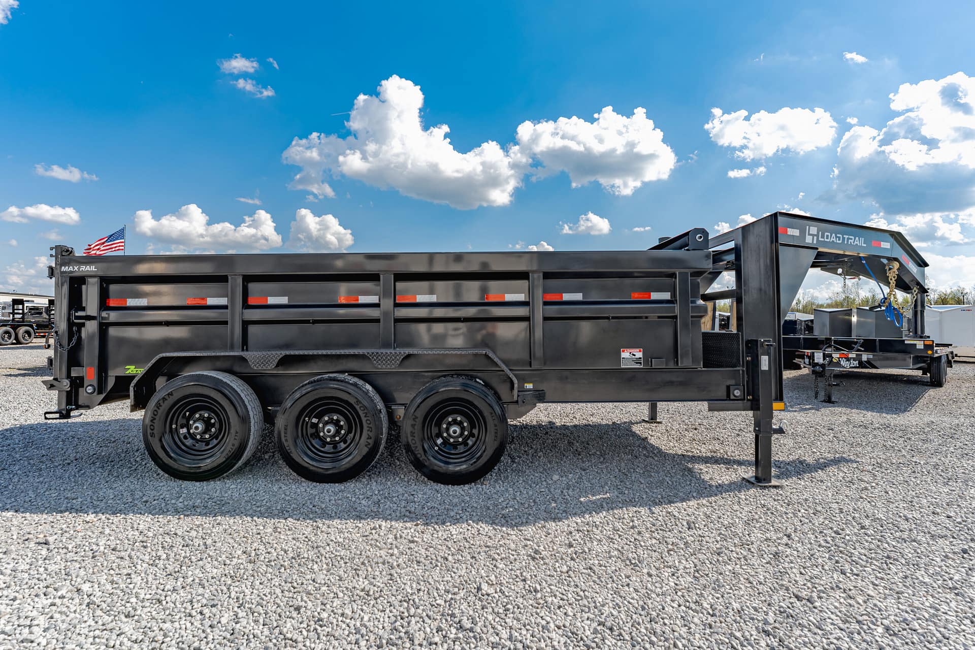 Black multi-axle dump trailer with red reflective stripes, parked on gravel under a blue sky; an American flag hangs from the front.